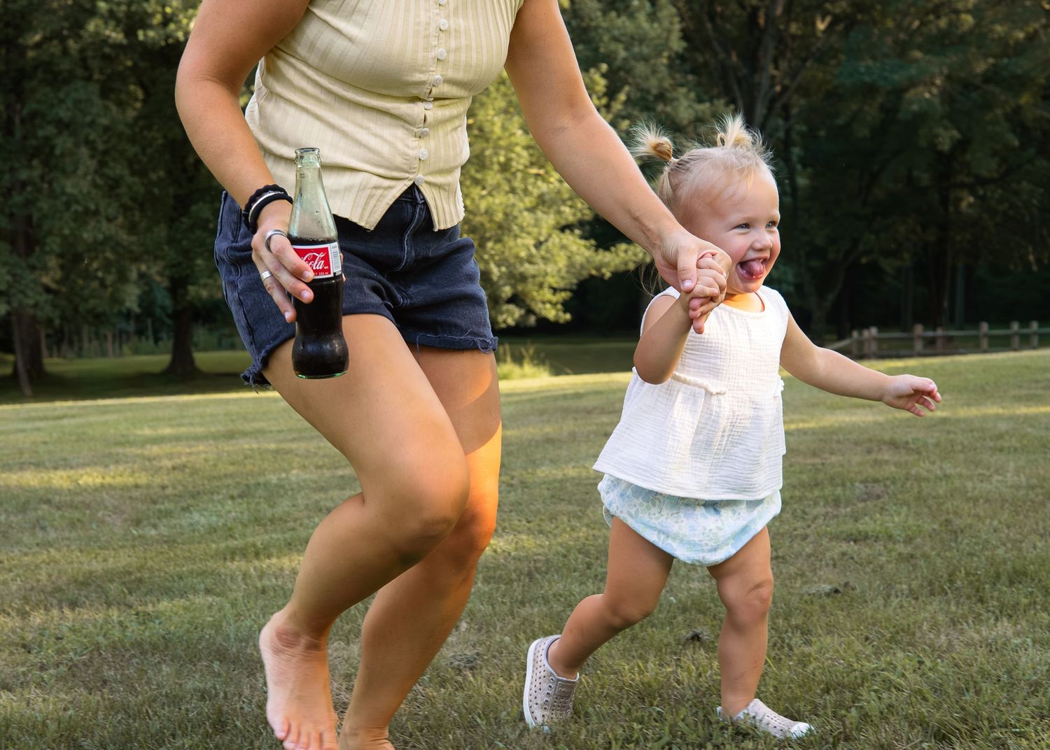 A mother holds a Coca-Cola bottle while joyfully running with her laughing toddler on a sunny green lawn.