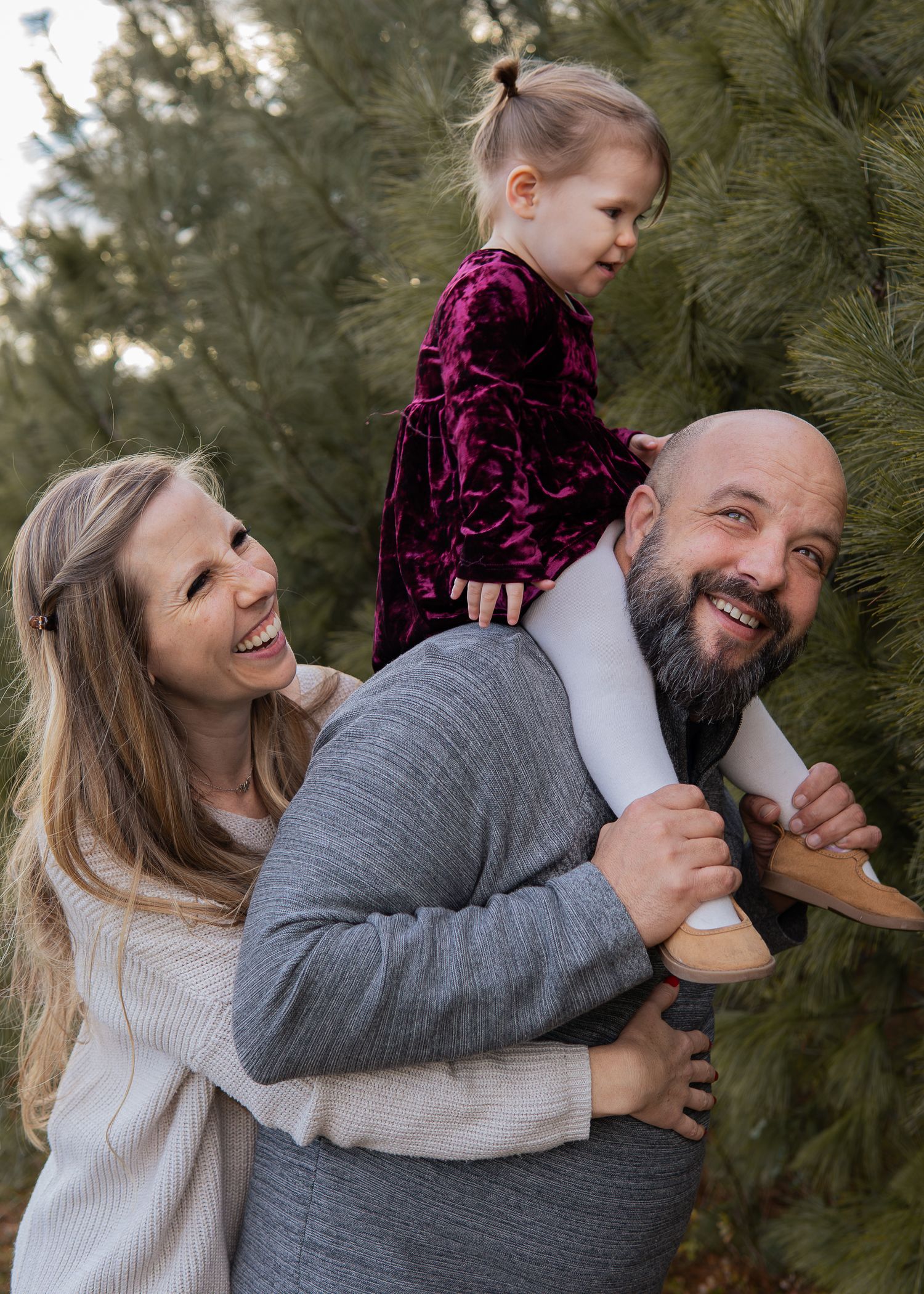 Laughing family with dad carrying young daughter on shoulders beside evergreen trees.