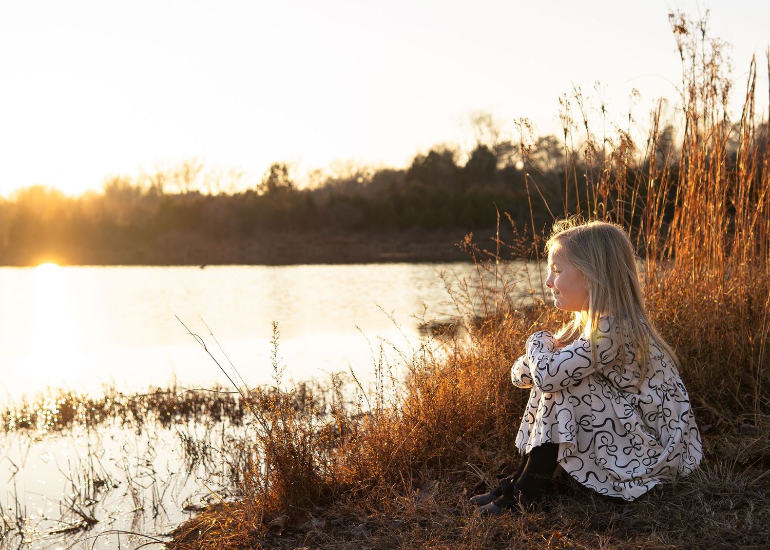 Young girl sitting by a sunlit lake at golden hour, surrounded by tall golden grass and autumn trees.