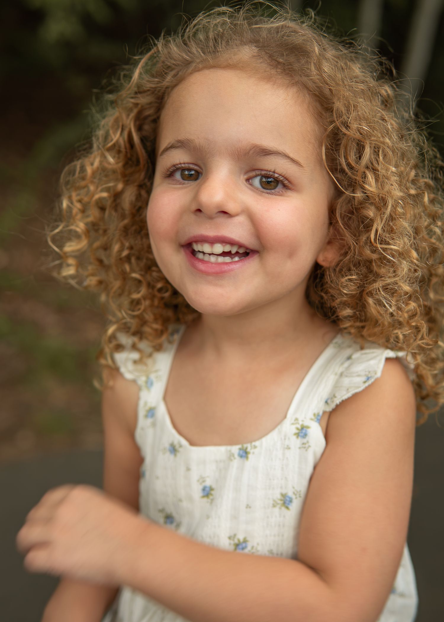 A smiling young girl with curly blonde hair wearing a floral white dress outdoors.