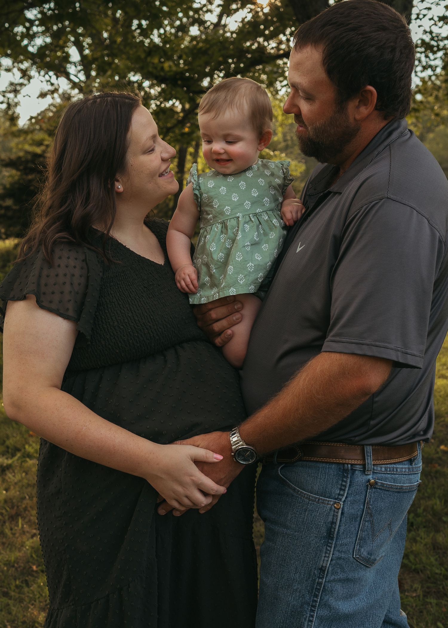 Pregnant woman and man holding smiling baby girl outdoors during golden hour family maternity photo session.