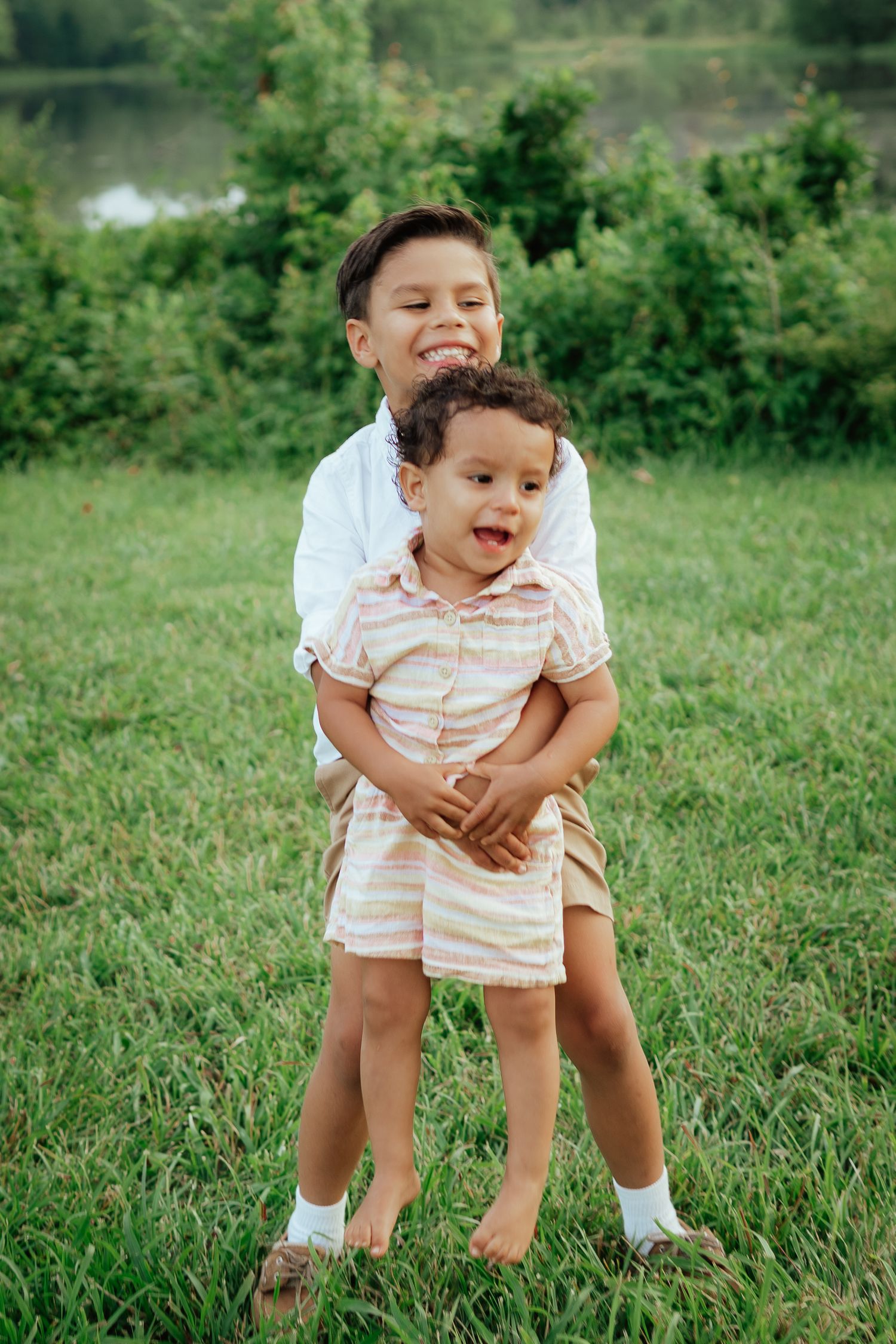 A heartwarming playful moment on grassy lawn with siblings hugging while standing near a lake and greenery.