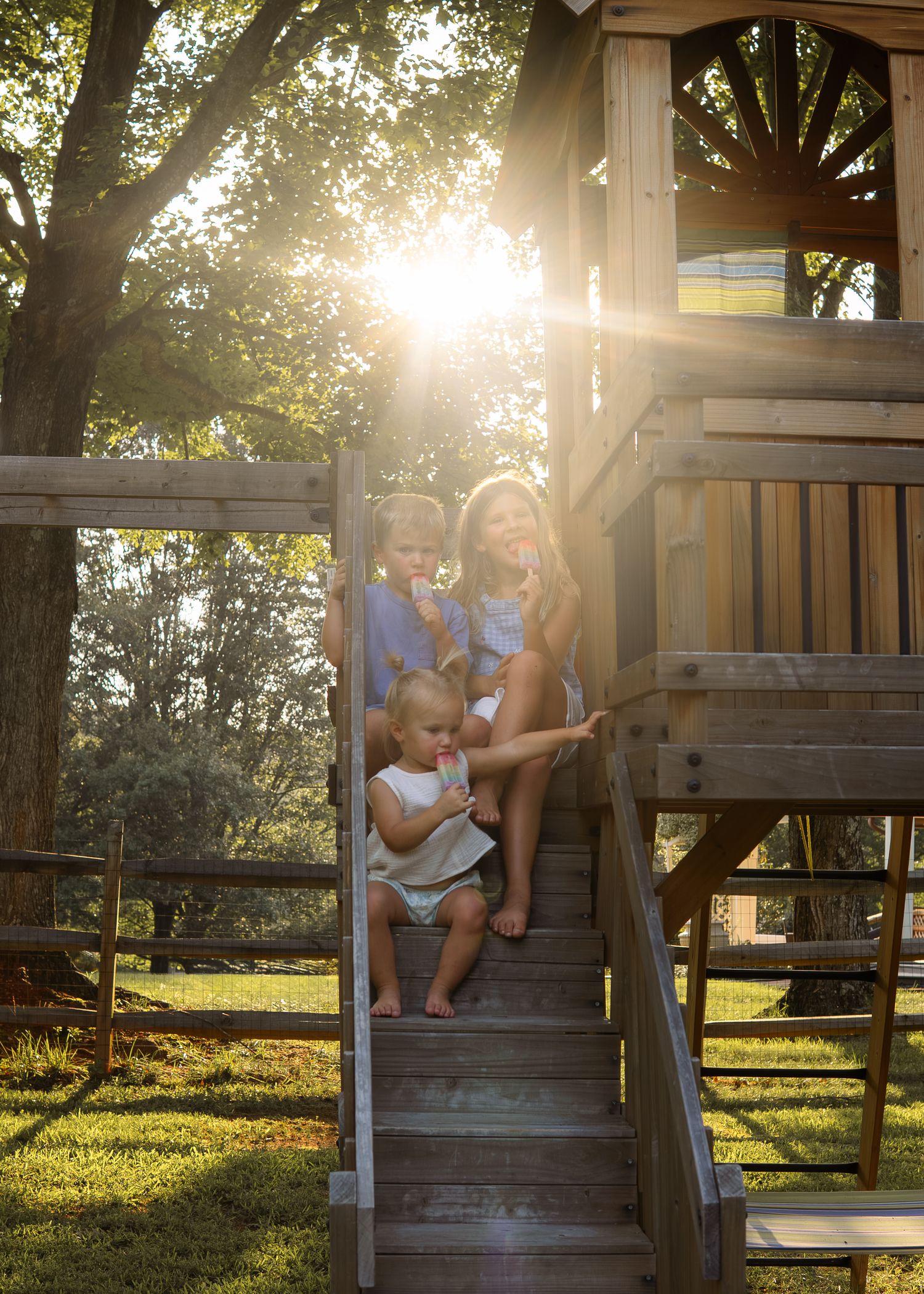 A mother and two young children sit on wooden treehouse steps, bathed in golden sunlight filtering through lush trees.