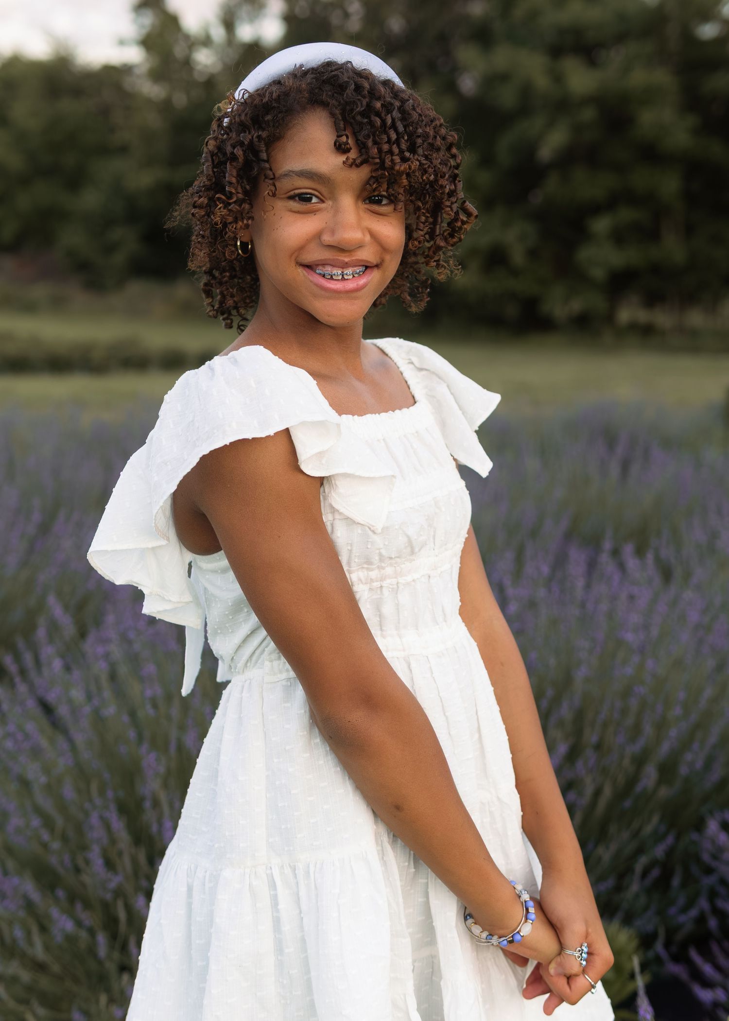 Young Black girl in white ruffled dress with headband smiling in lavender field at sunset.
