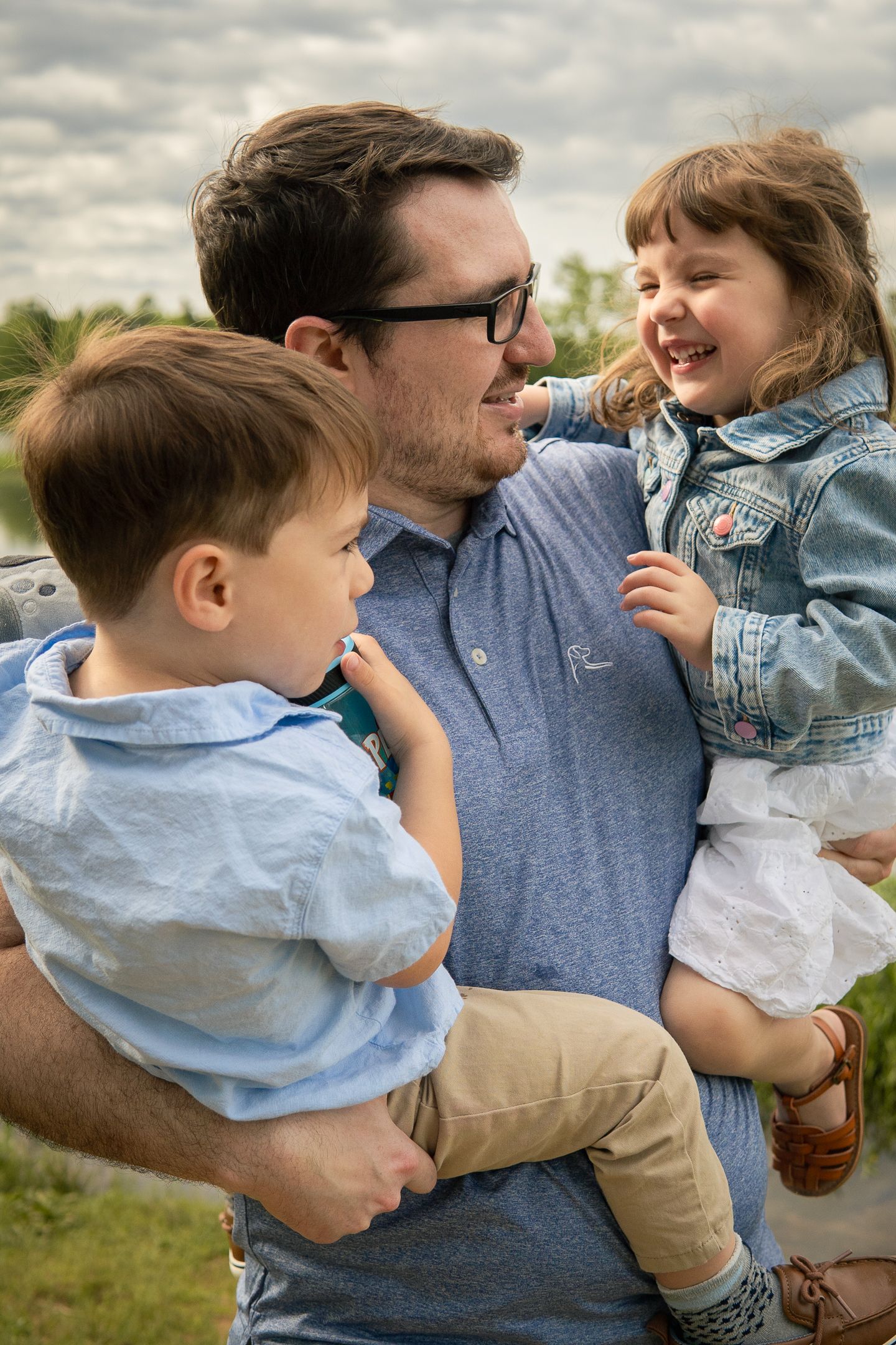 Joyful outdoor family moment with parent holding two young children in casual attire against cloudy sky.