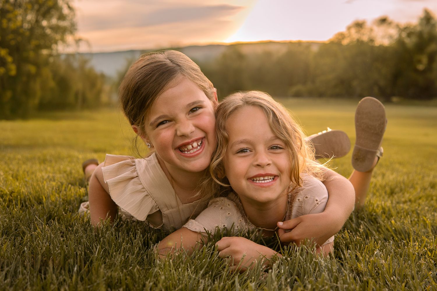 Two children lying together in a grassy field at sunset, laughing joyfully with warm golden light behind them.
