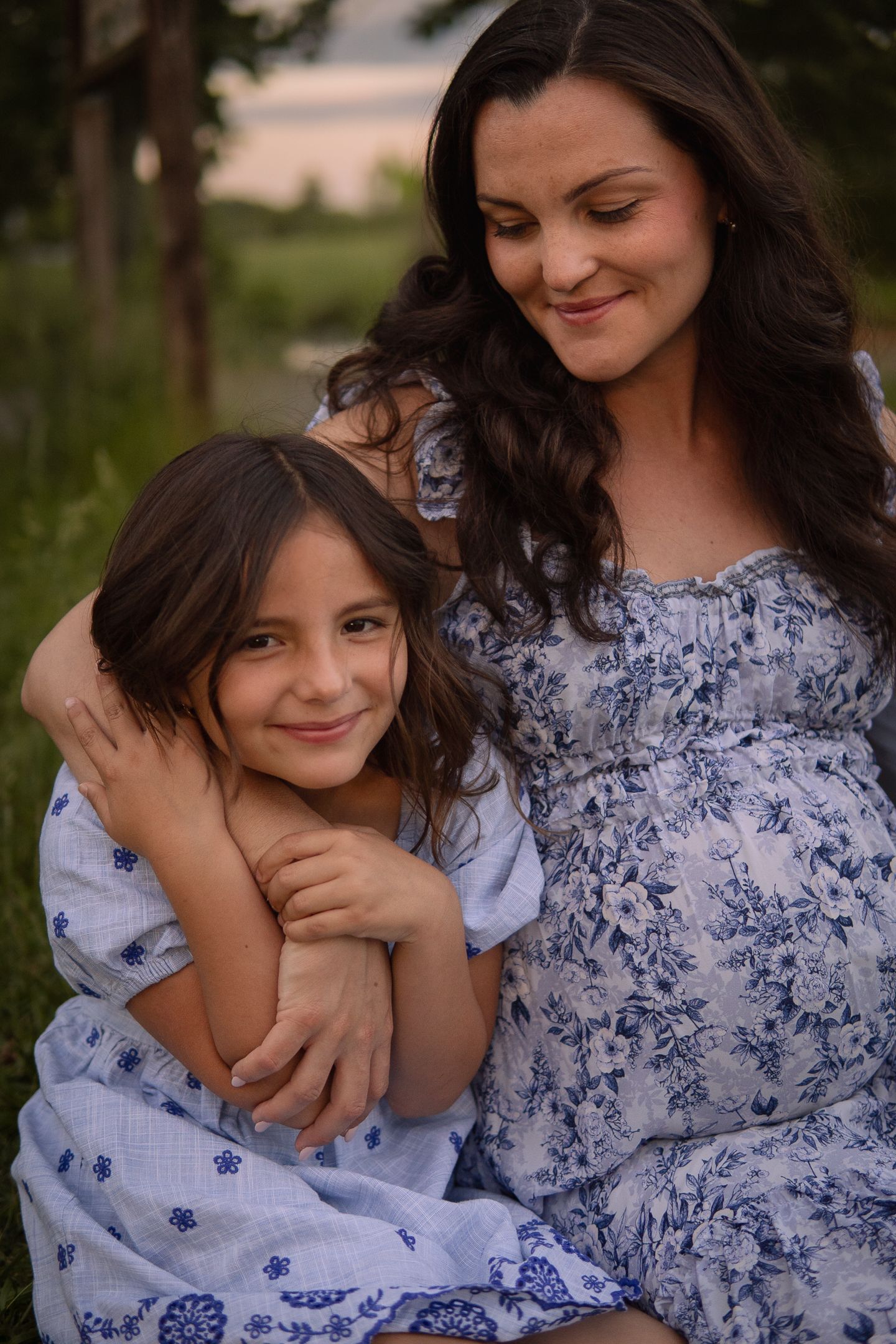 A mother and daughter in coordinating blue and white dresses share an intimate embrace during an outdoor photo session.