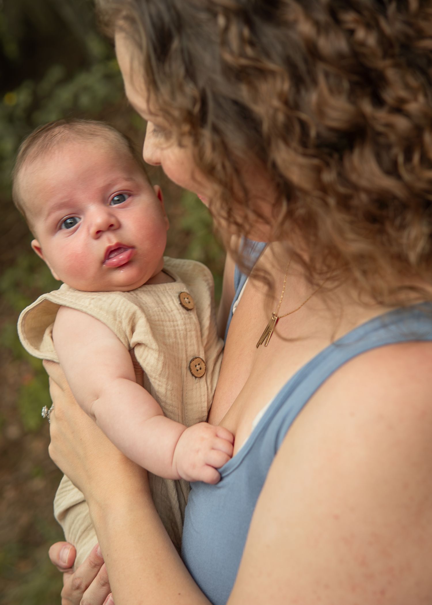 A curly-haired woman in a blue top holds a wide-eyed baby in a tan romper outdoors.