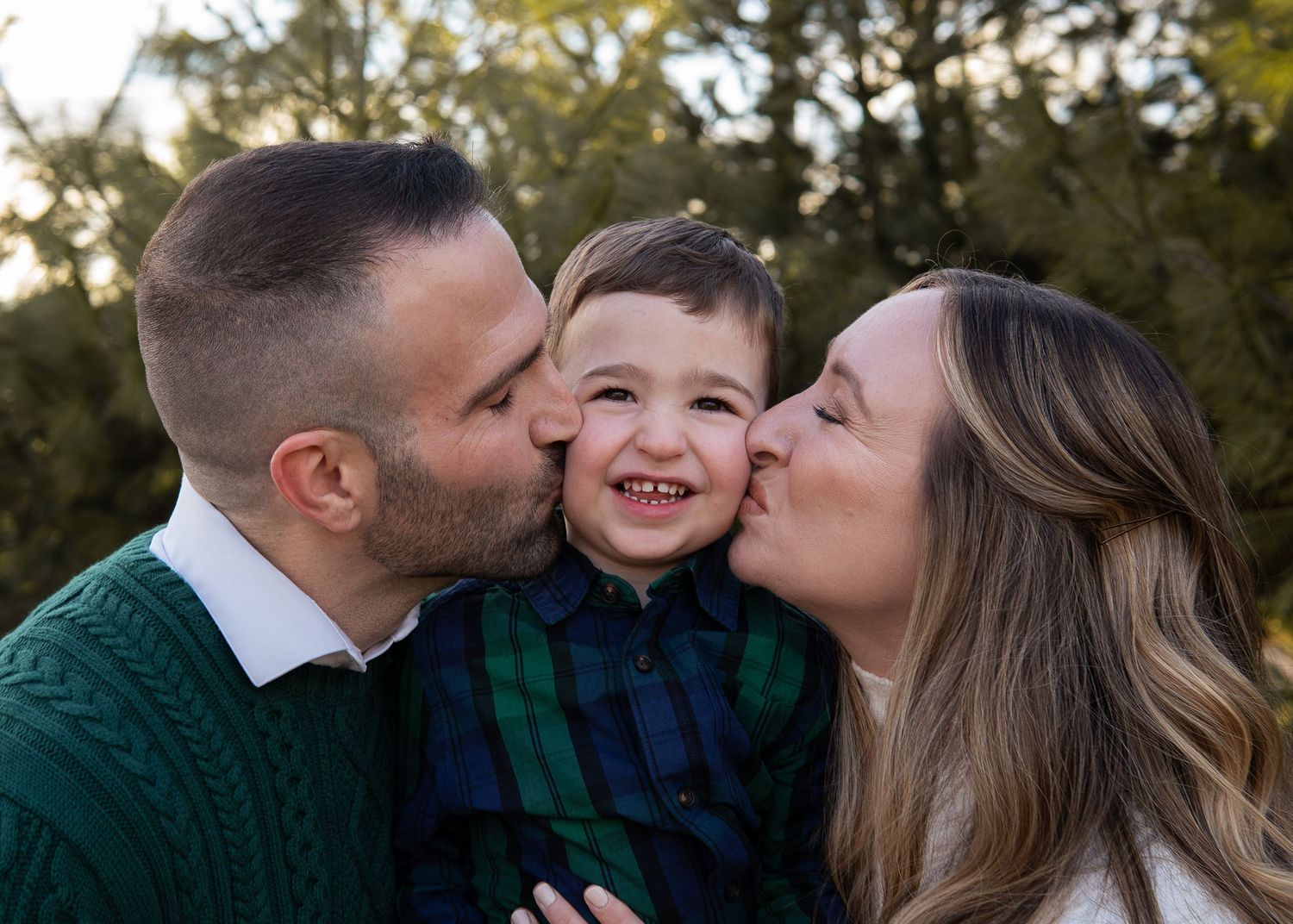 Happy toddler boy smiling while parents kiss his cheeks outdoors in a family portrait session.