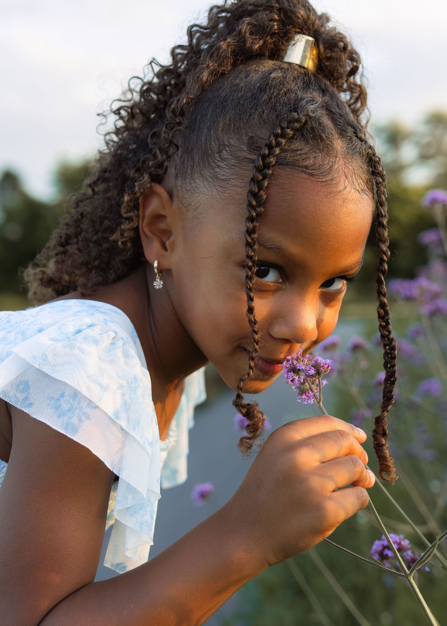 Young Black girl with braided hair smelling purple wildflowers in a garden at golden hour.