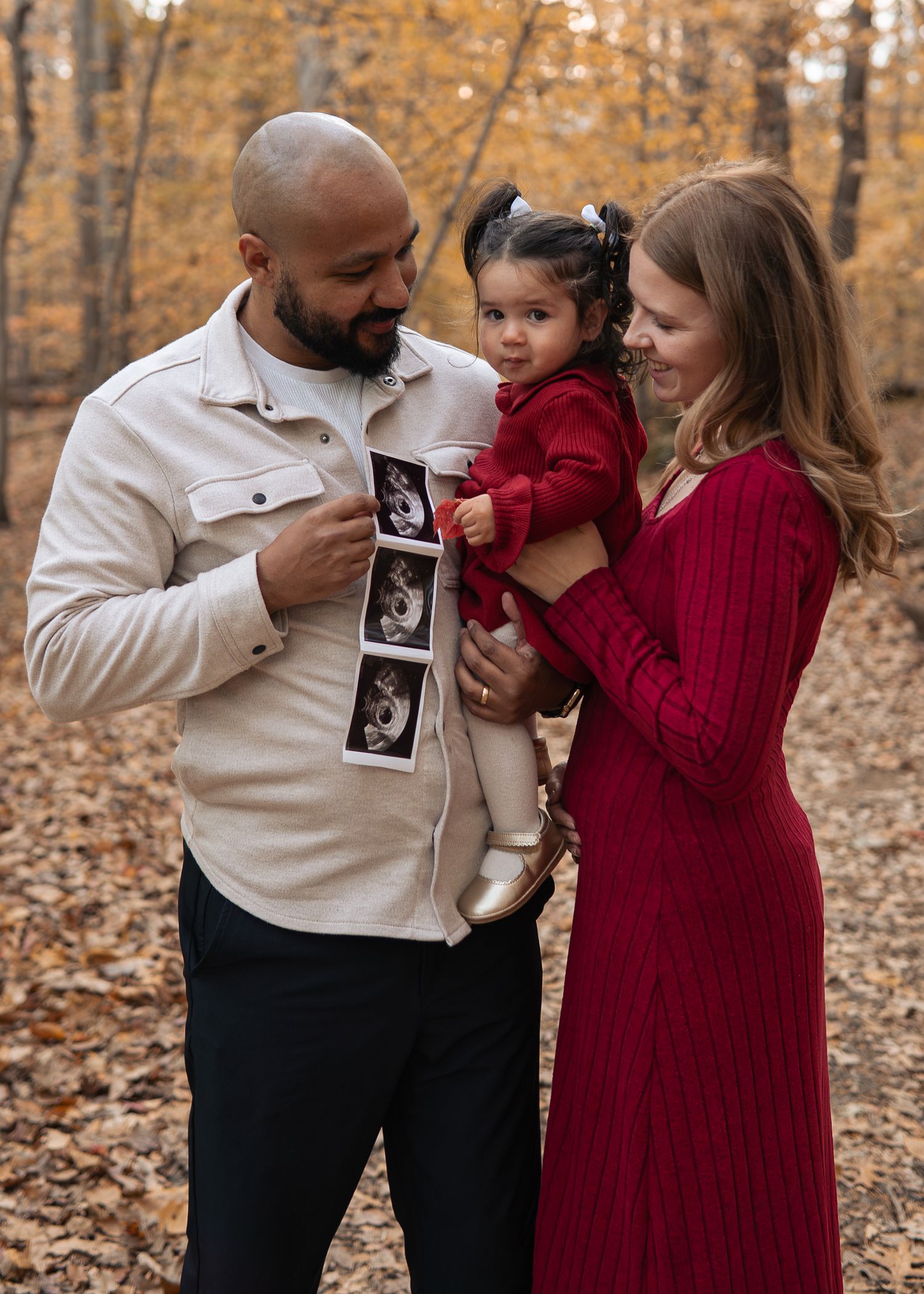 Family of three in autumn woods, dad holding ultrasound photos, mom in red dress holding toddler girl.