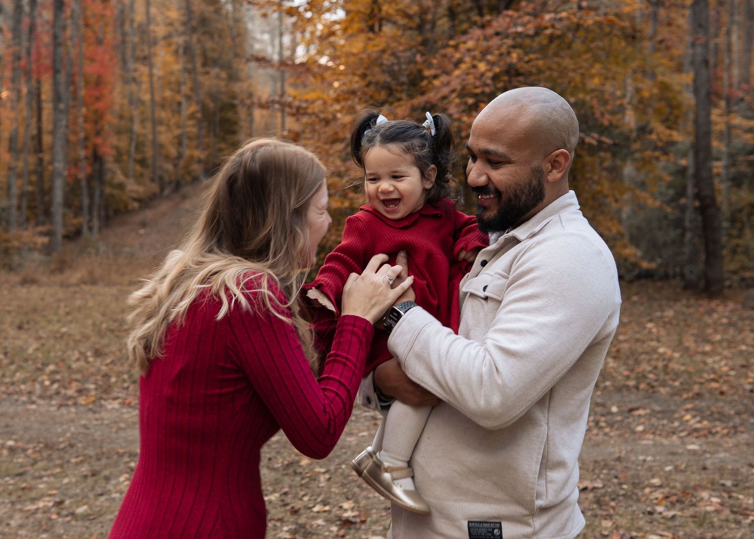 Family of three enjoying autumn outdoors, father holding toddler girl in red dress while mother smiles.