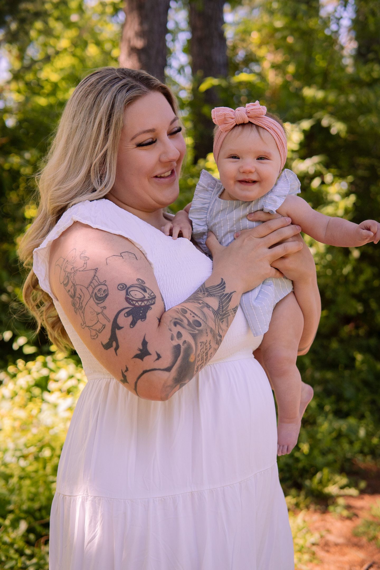 A parent with decorative arm tattoos holds a smiling baby wearing a pink bow in an outdoor setting.