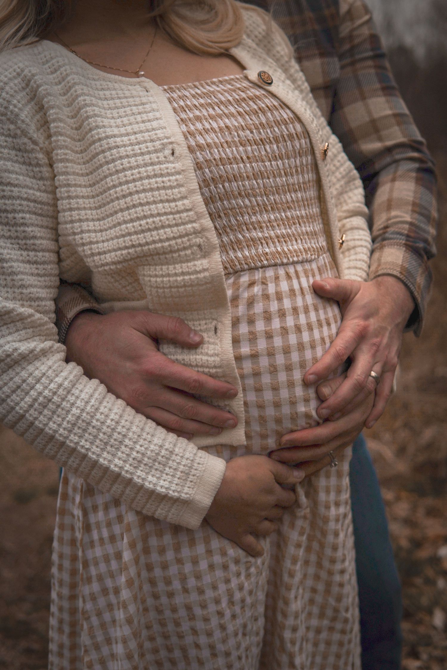 Close-up of hands cradling a pregnant belly wearing a cream knit cardigan and gingham dress in an autumn outdoor setting.