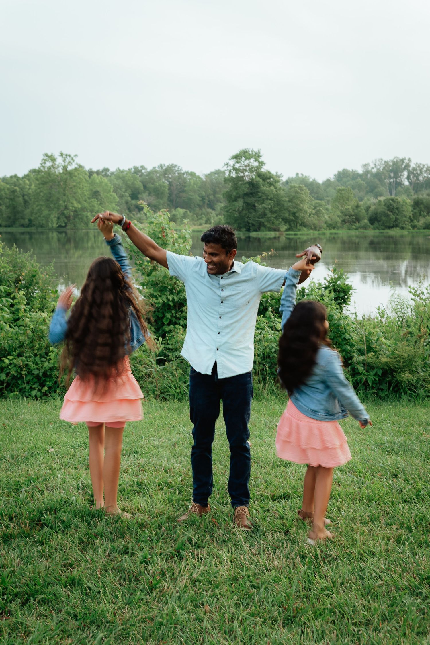 A family plays and dances together in a grassy field by a lake on a sunny day.
