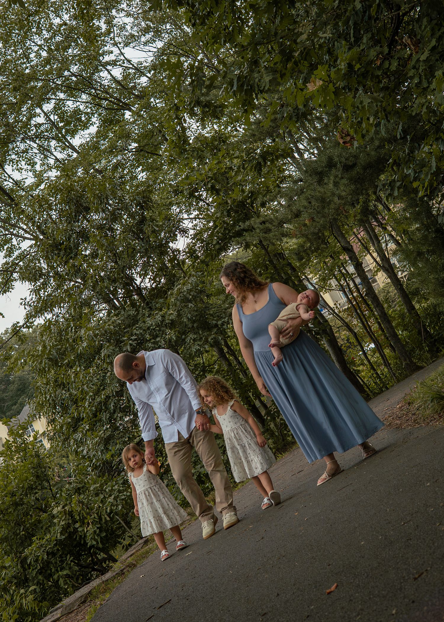 Family of five walks along a tree-lined path, parents holding hands with toddlers as mother carries newborn in blue dress.