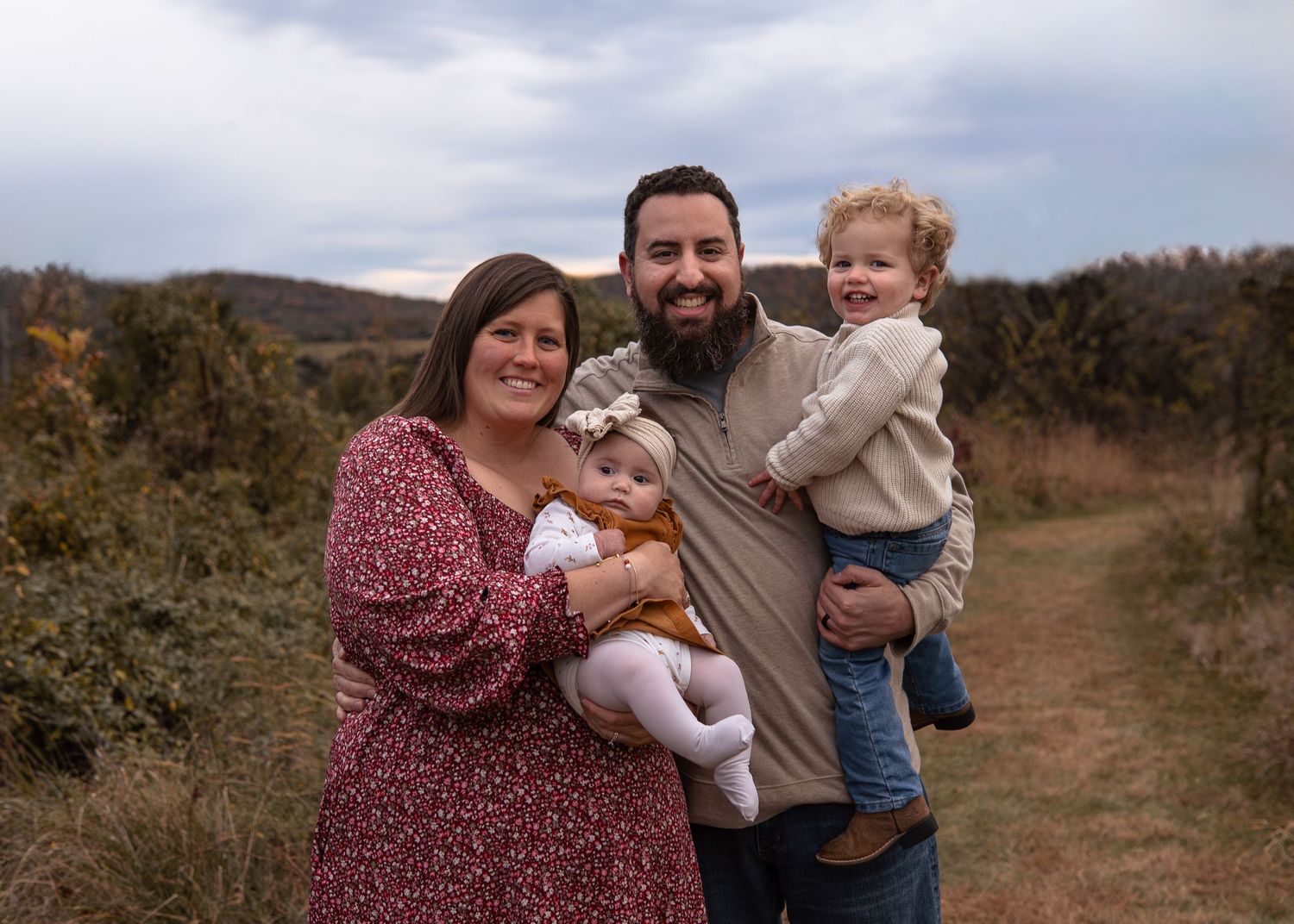 Family of four posing outdoors on a scenic trail, parents holding two young children, with rolling hills in the background.
