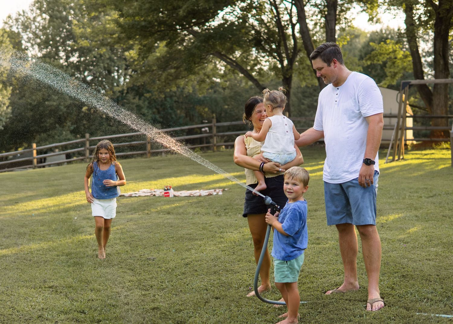 Family of five enjoying summer fun with a garden hose, spraying water on a sunny green lawn.
