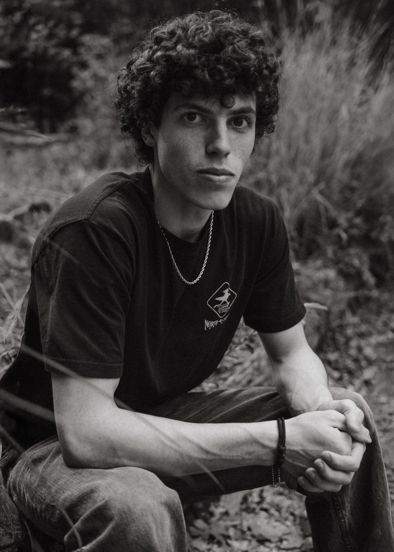 Black and white portrait of a young man with curly hair wearing a black t-shirt and chain necklace outdoors.
