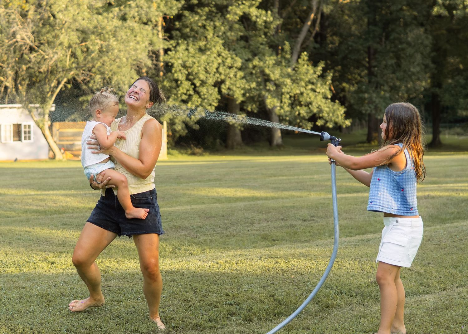 Woman holding baby laughs as girl sprays water hose at her in sunny backyard.