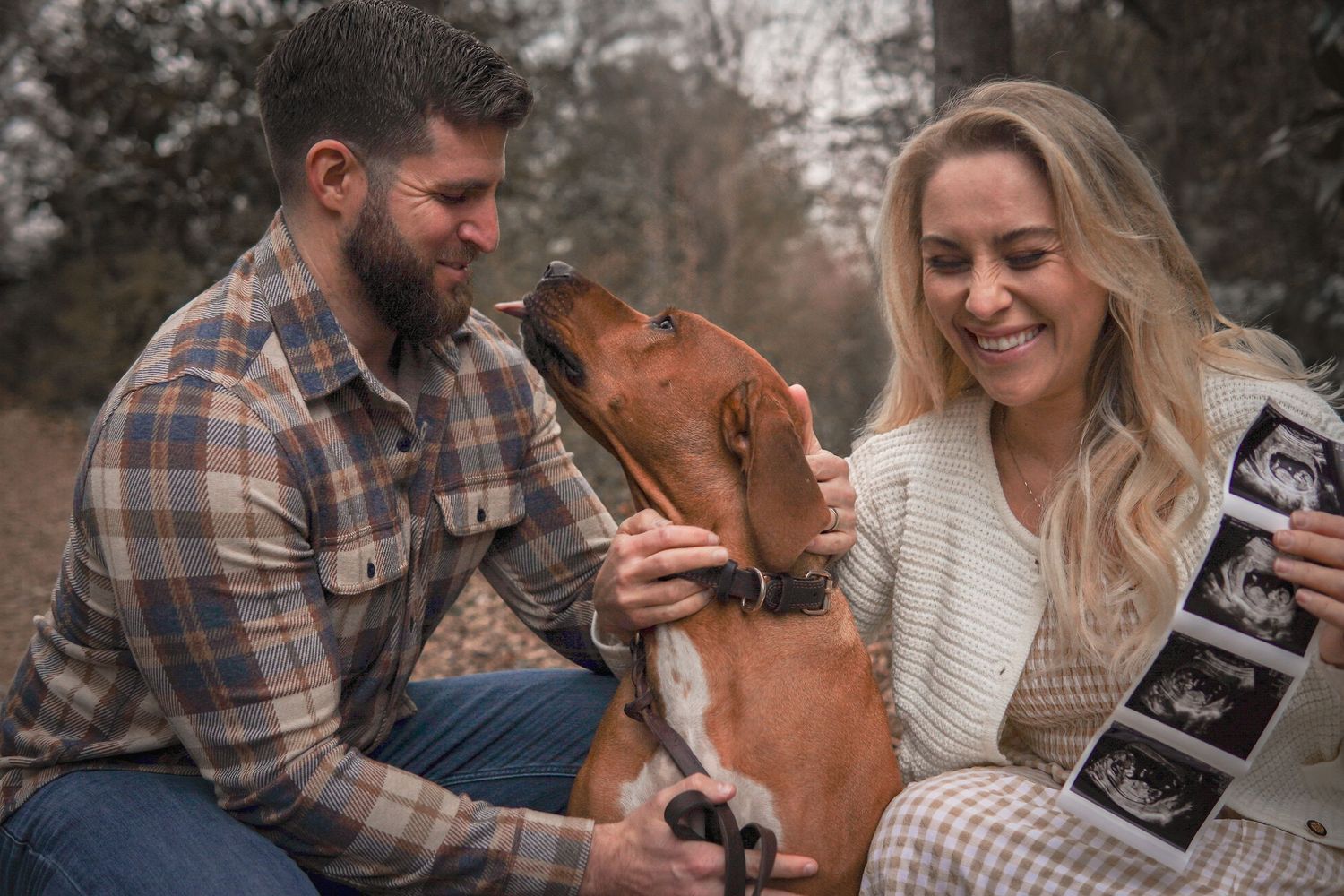 A couple sits together with their dog in nature while displaying ultrasound images for a pregnancy announcement.