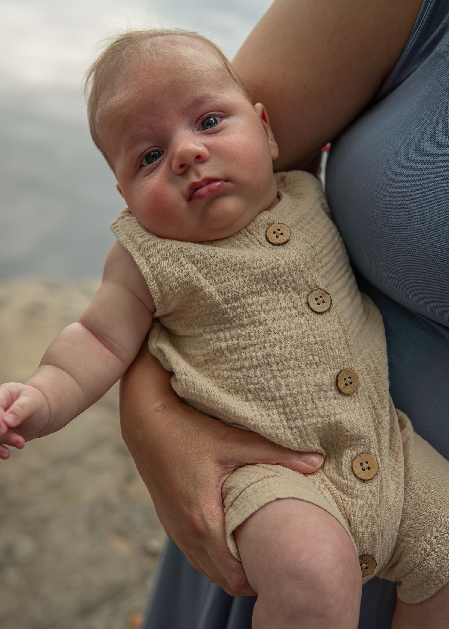 Adorable newborn baby in a beige linen romper with buttons being held by a woman in a gray dress.