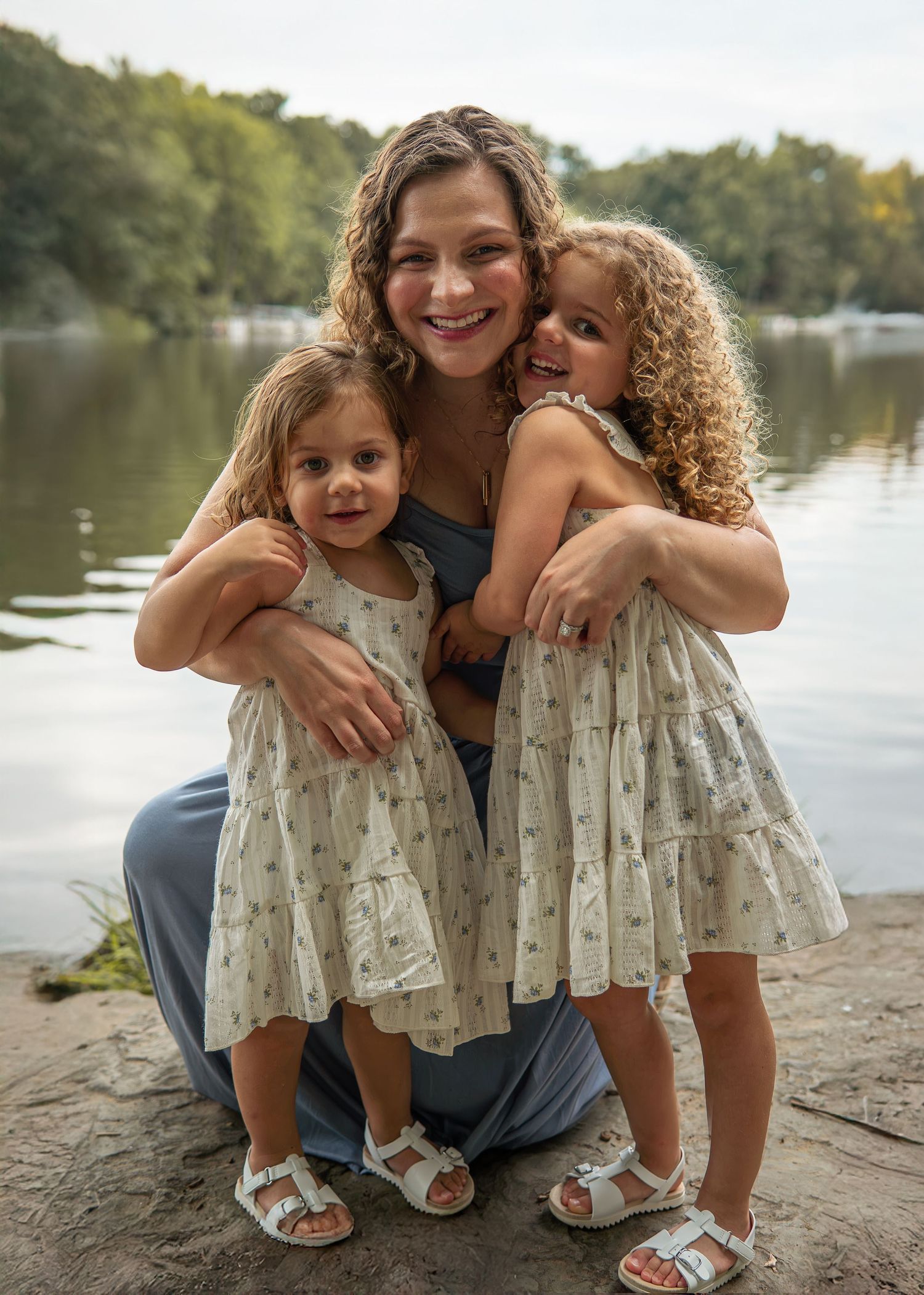 A smiling woman crouches by a lake hugging two young girls in matching floral dresses and white sandals outdoors.