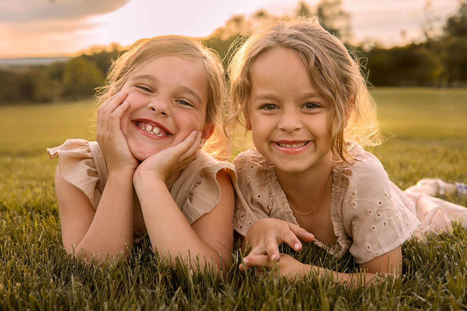 Two young children lying together in a grassy field at sunset, laughing playfully at the camera.