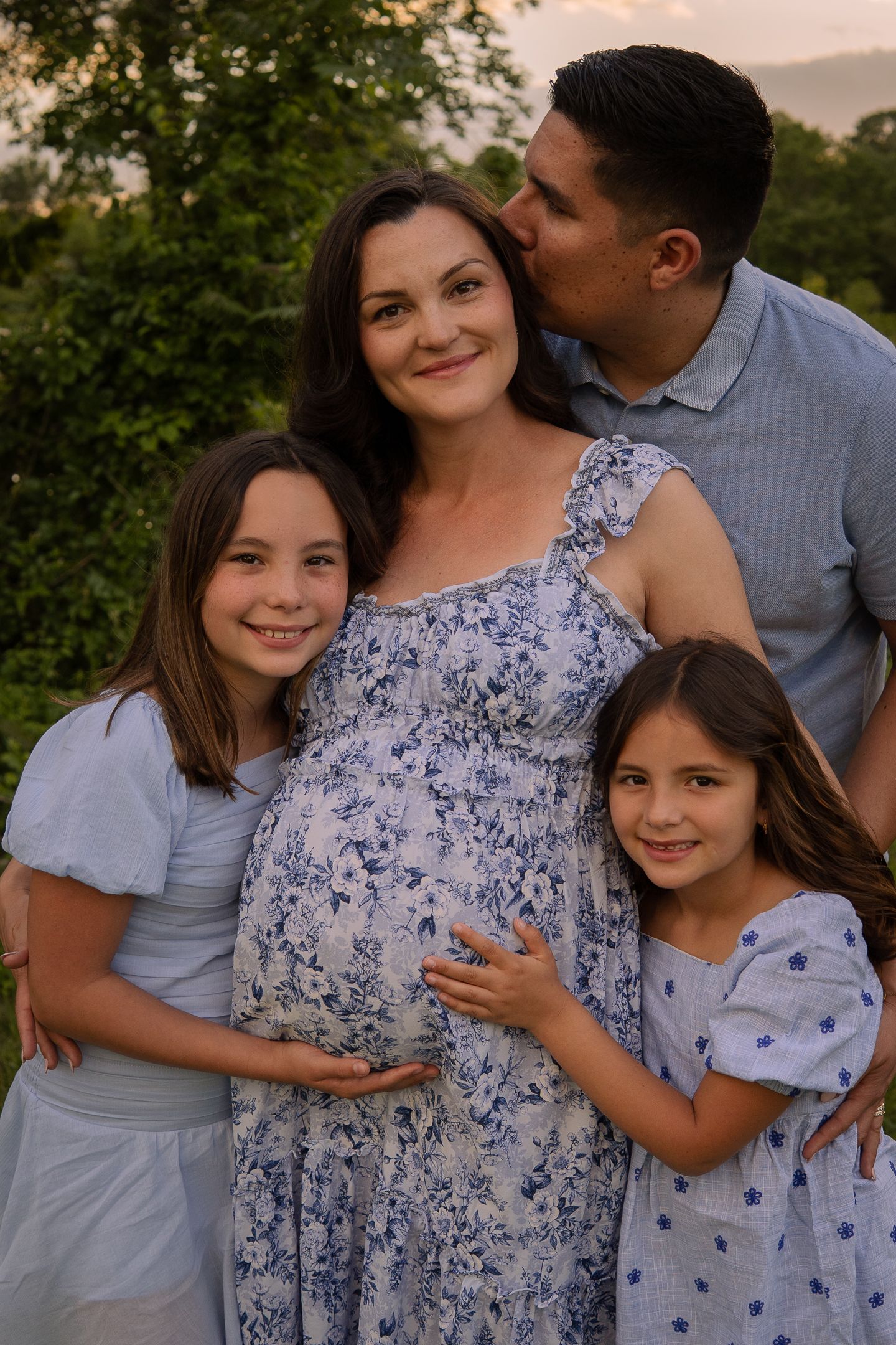 A tender family maternity photo outdoors at sunset with coordinated blue and white outfits.