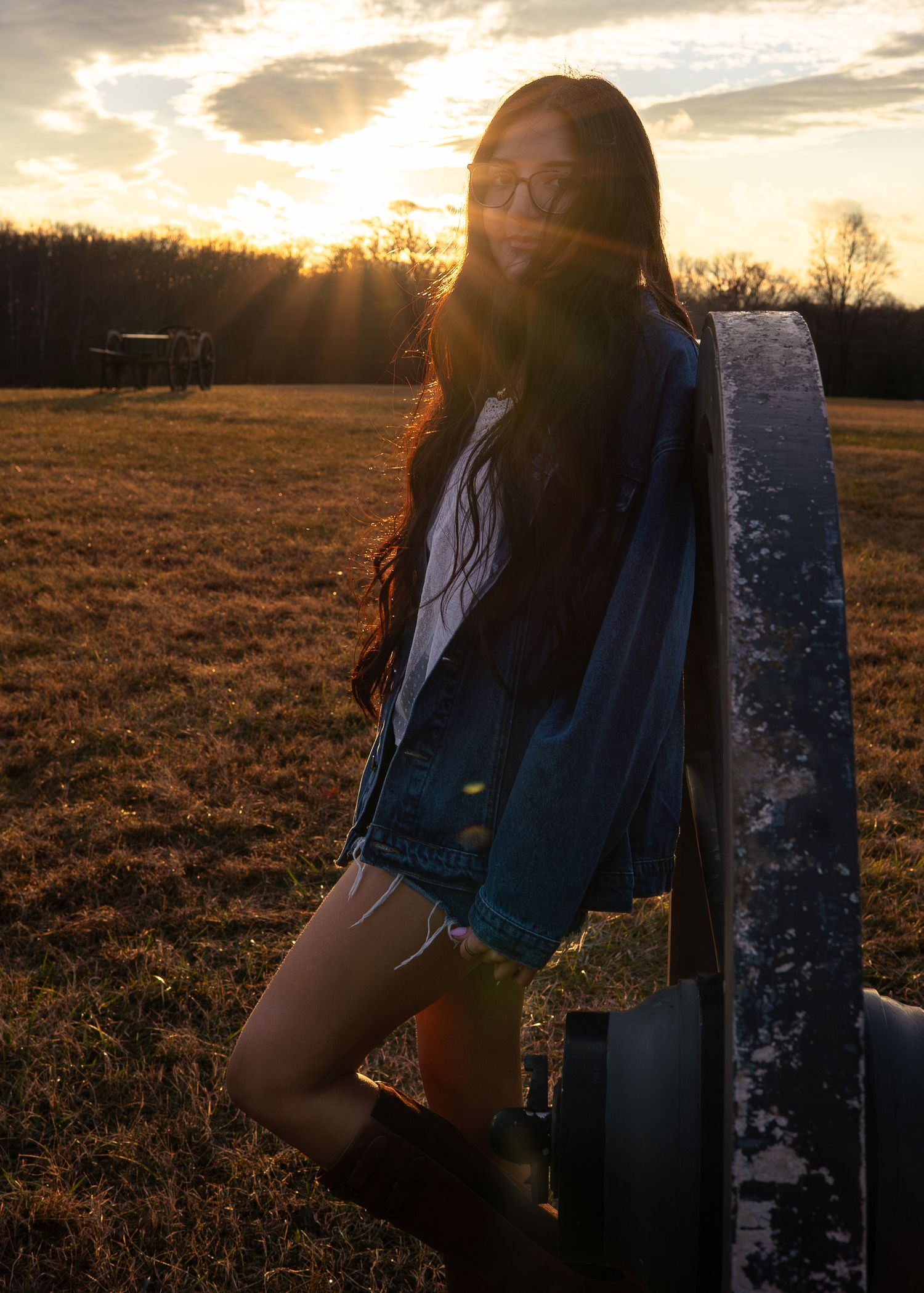 Young woman in denim jacket and shorts leaning on post in field at dramatic sunset.