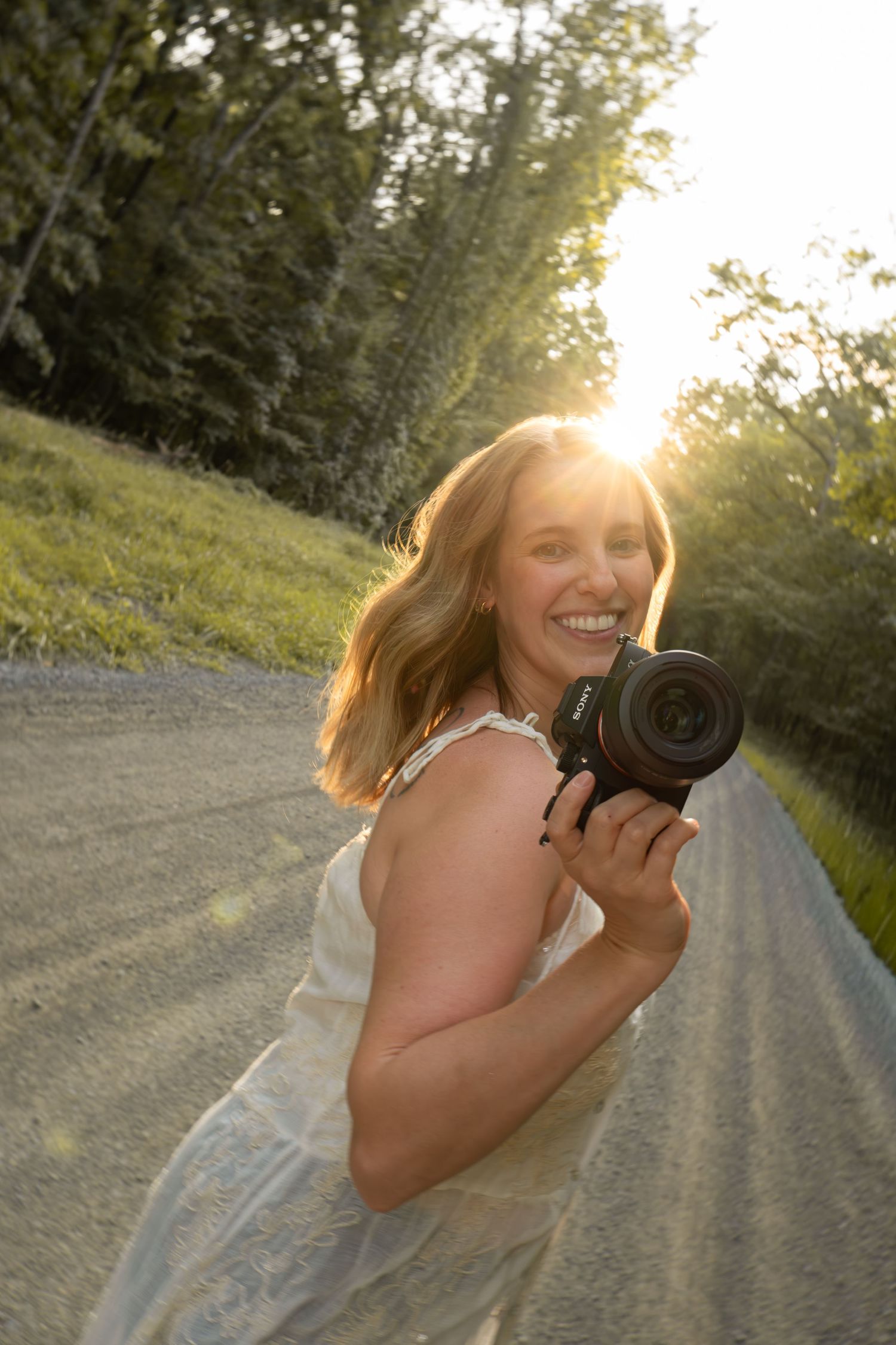 Smiling woman holding a DSLR camera on a sunlit gravel road surrounded by lush green trees at golden hour.