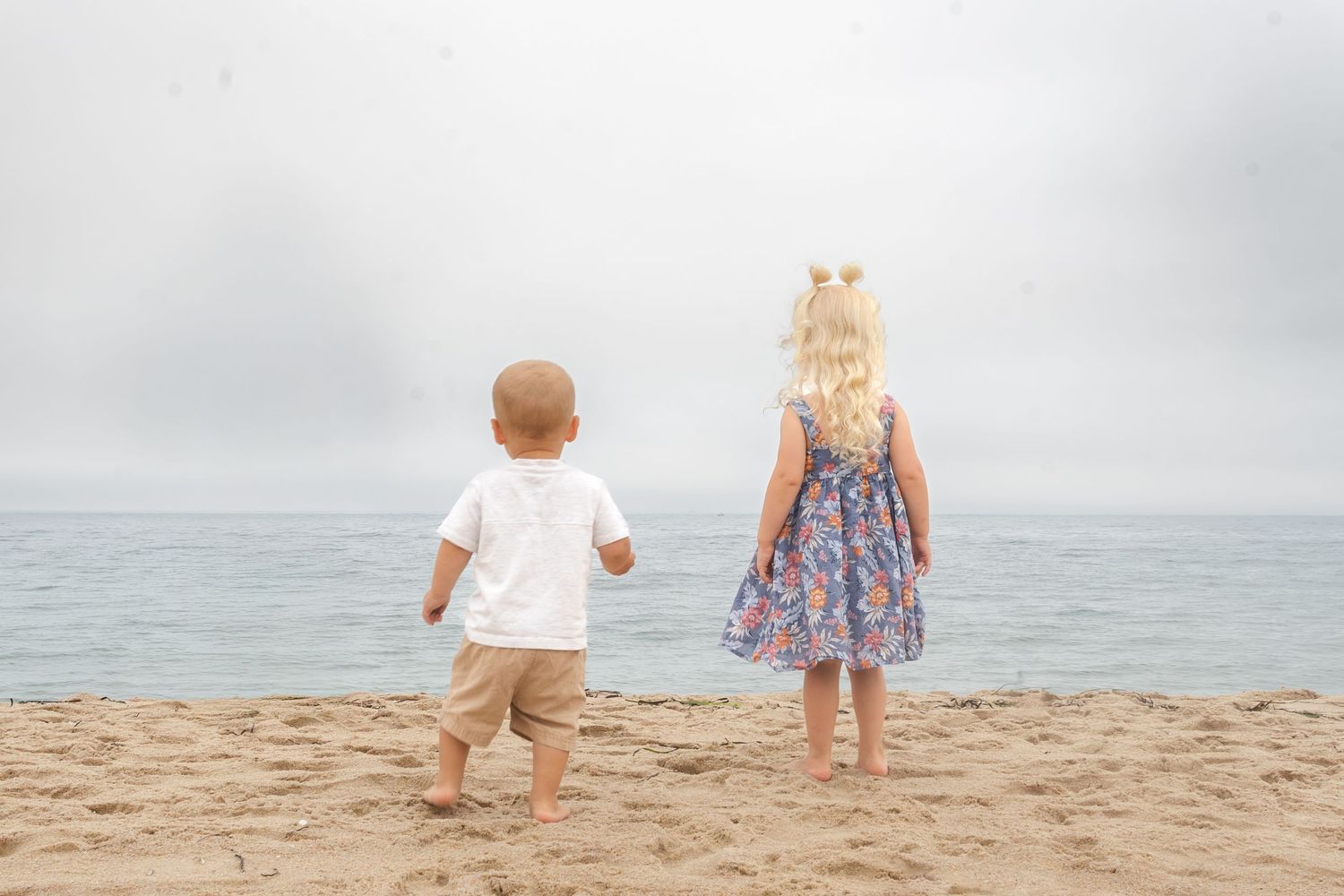 Two young children standing on a sandy beach gazing at the calm ocean on an overcast day.