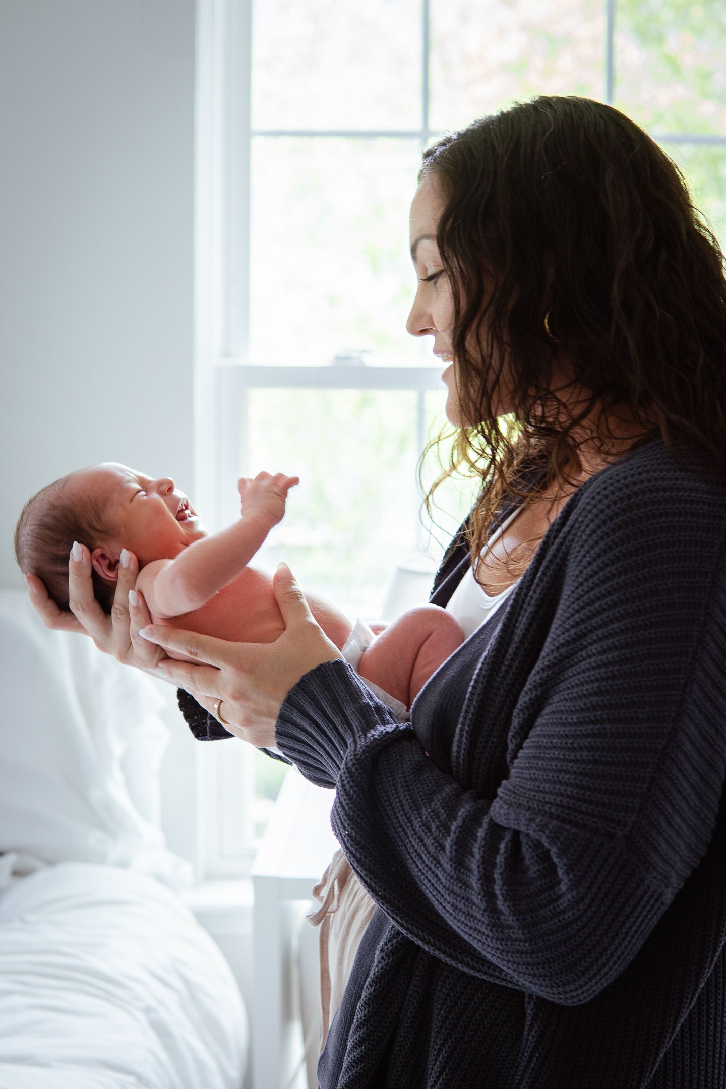 A parent in a navy sweater holds and gazes at their newborn by a bright window.