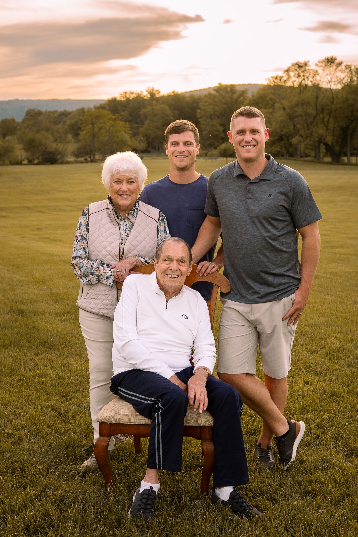 Four generations gather for an intimate outdoor family portrait at sunset in a grassy field.