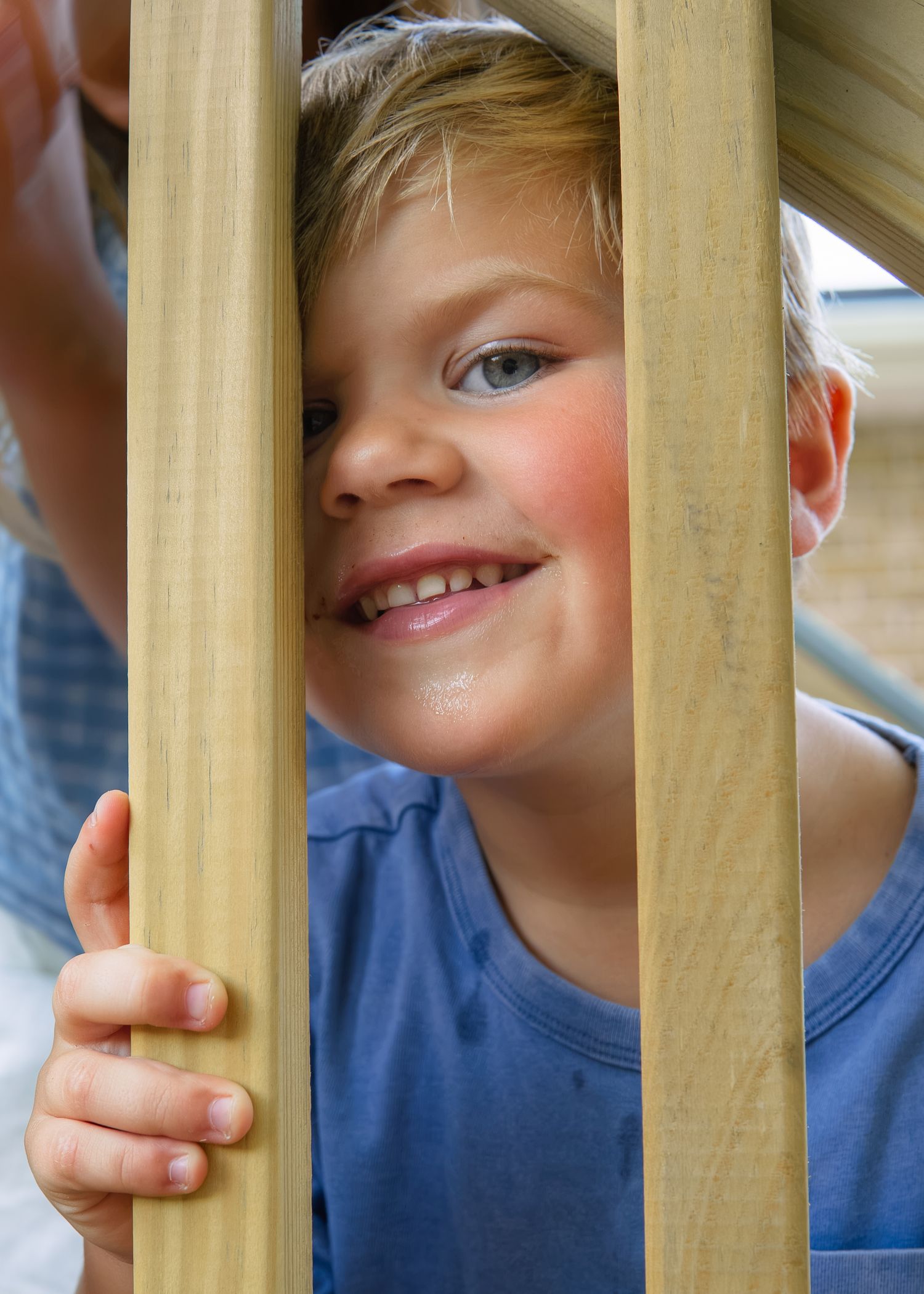 Smiling blonde boy peering through wooden playground railings on sunny day.