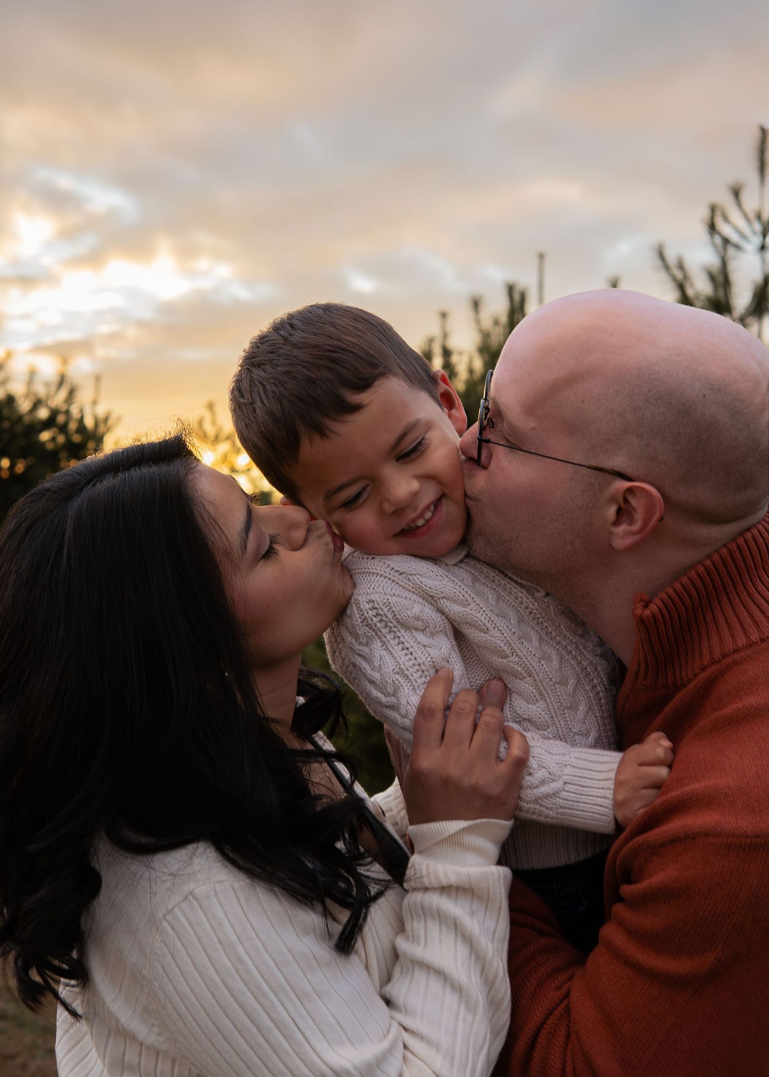 Parents kissing their smiling young son on both cheeks at sunset outdoors near evergreen trees.