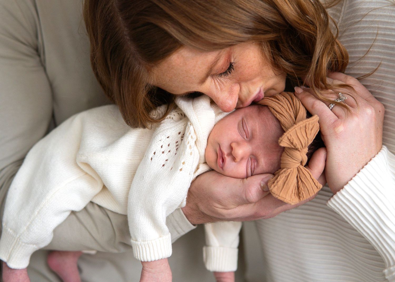 New mother tenderly kissing her sleeping newborn baby wearing a tan bow headband.