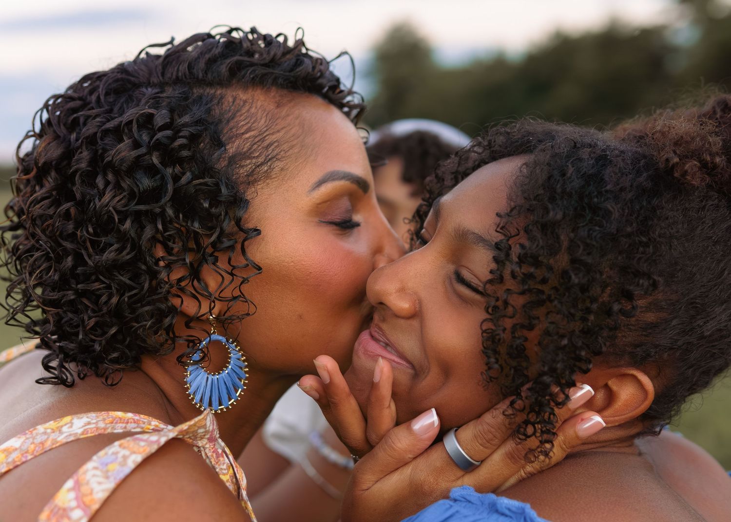 A mother shares a tender kiss on the cheek with her young daughter outdoors.