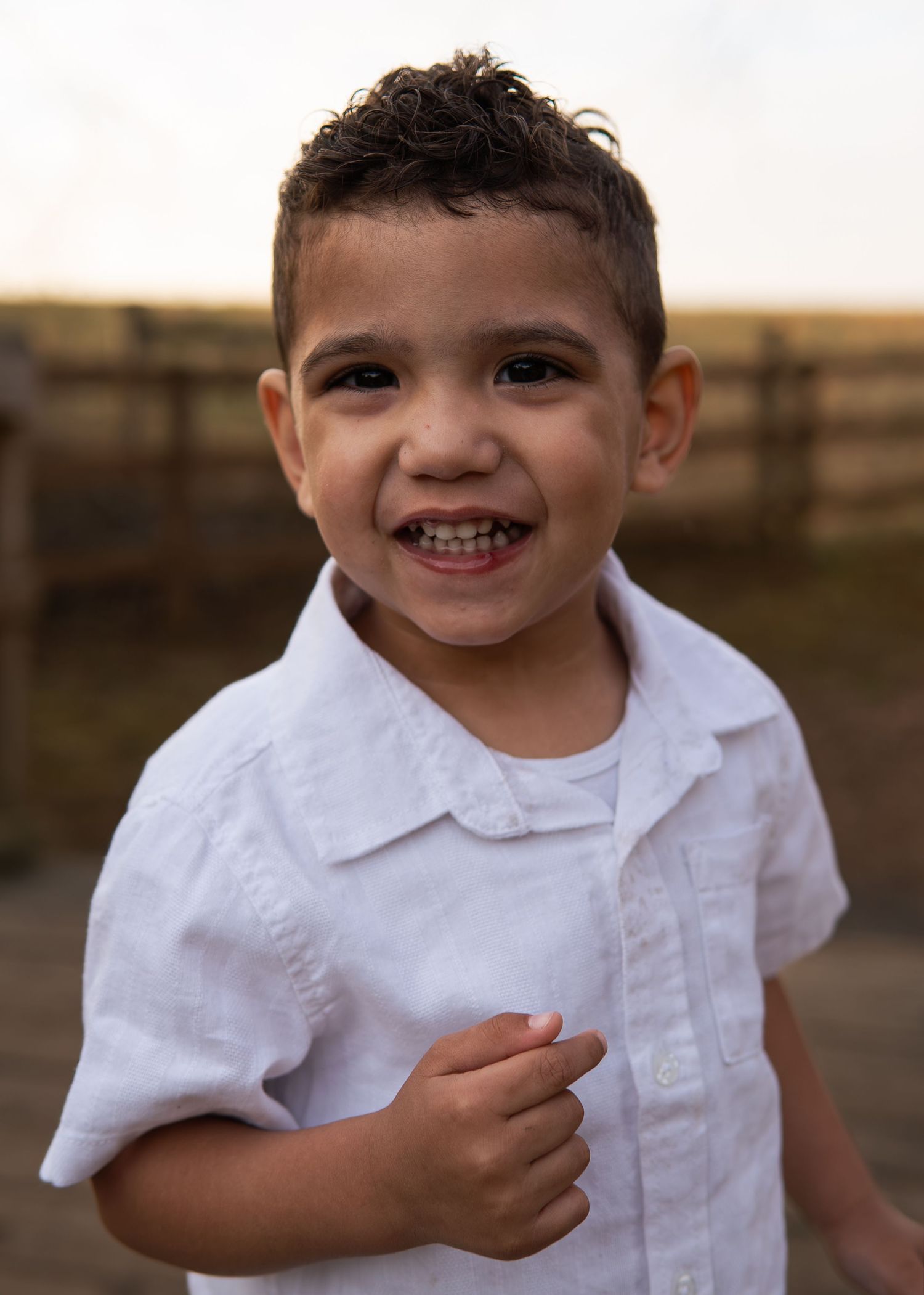 Happy young Hispanic boy in white button-down shirt smiling outdoors at golden hour near a fence.