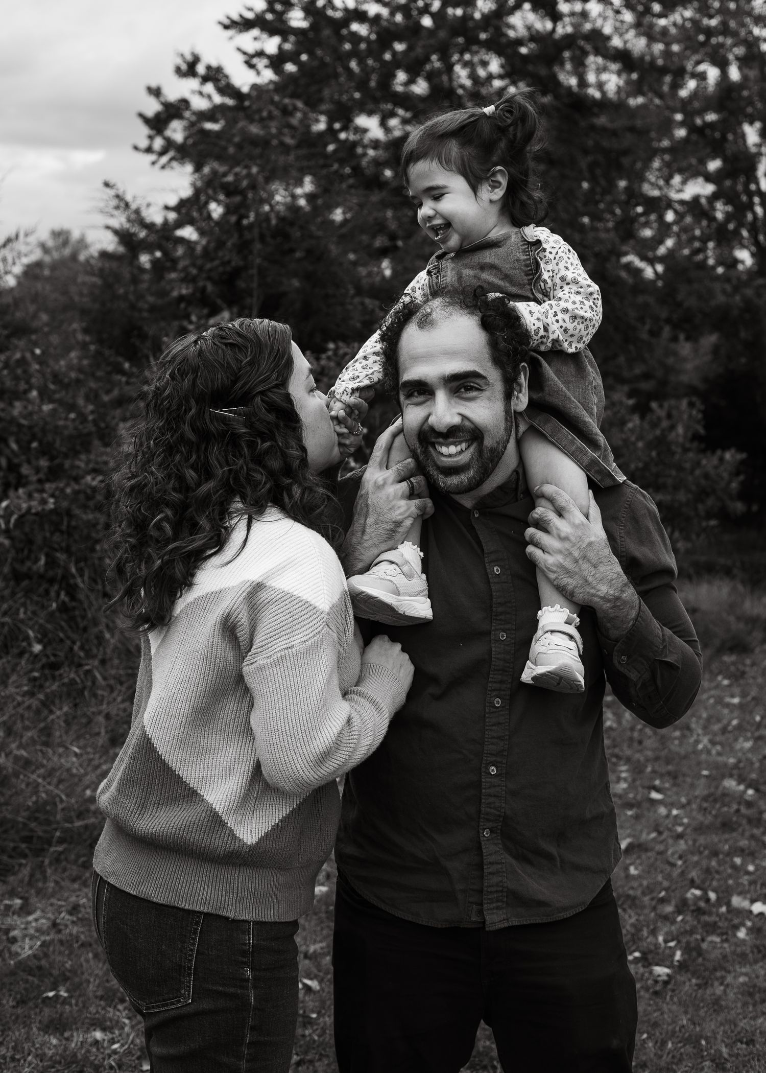 Black and white photo of joyful family with mother and father laughing as toddler sits on dads shoulders.