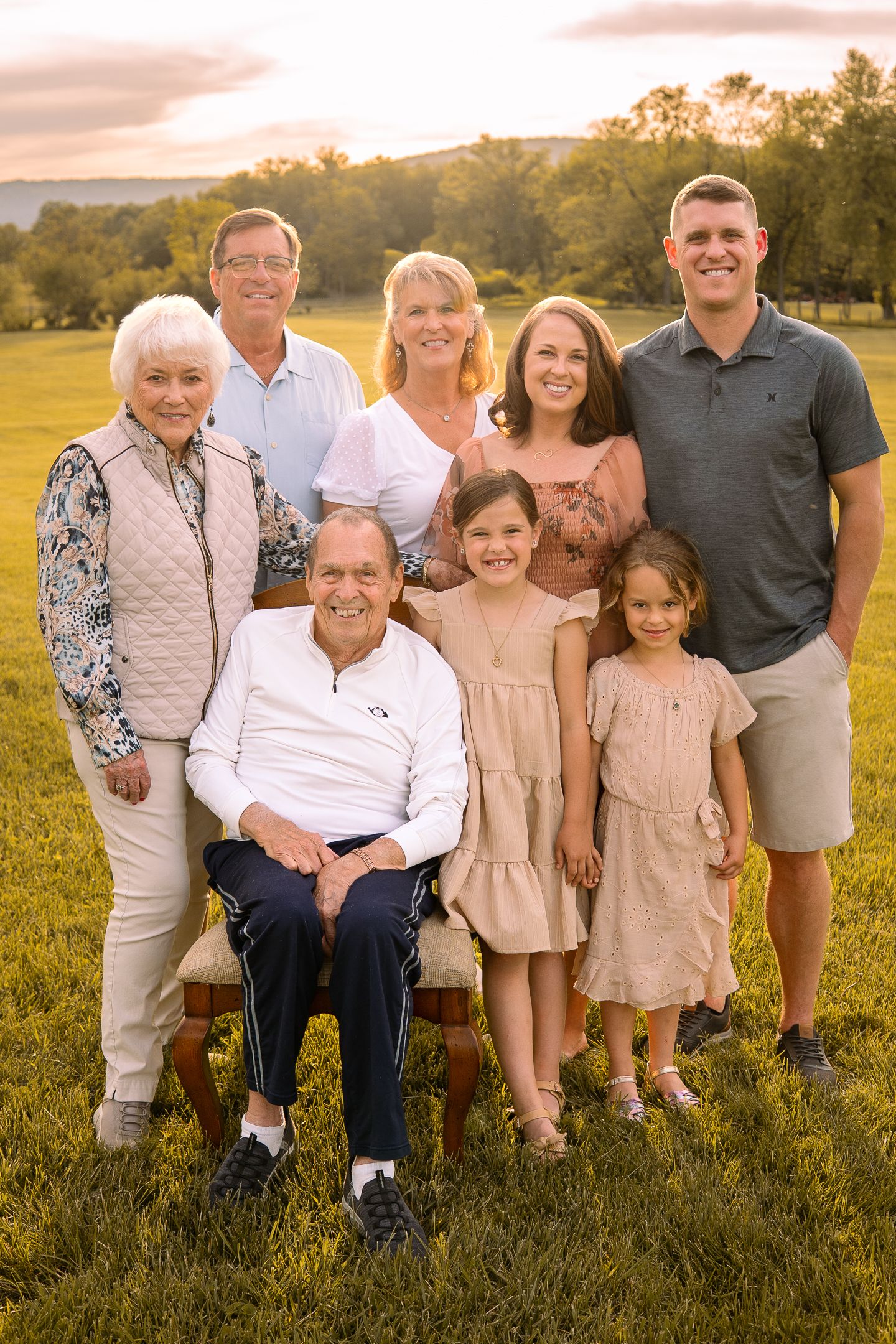 A multi-generational family portrait outdoors on a grassy hill during golden hour sunset.