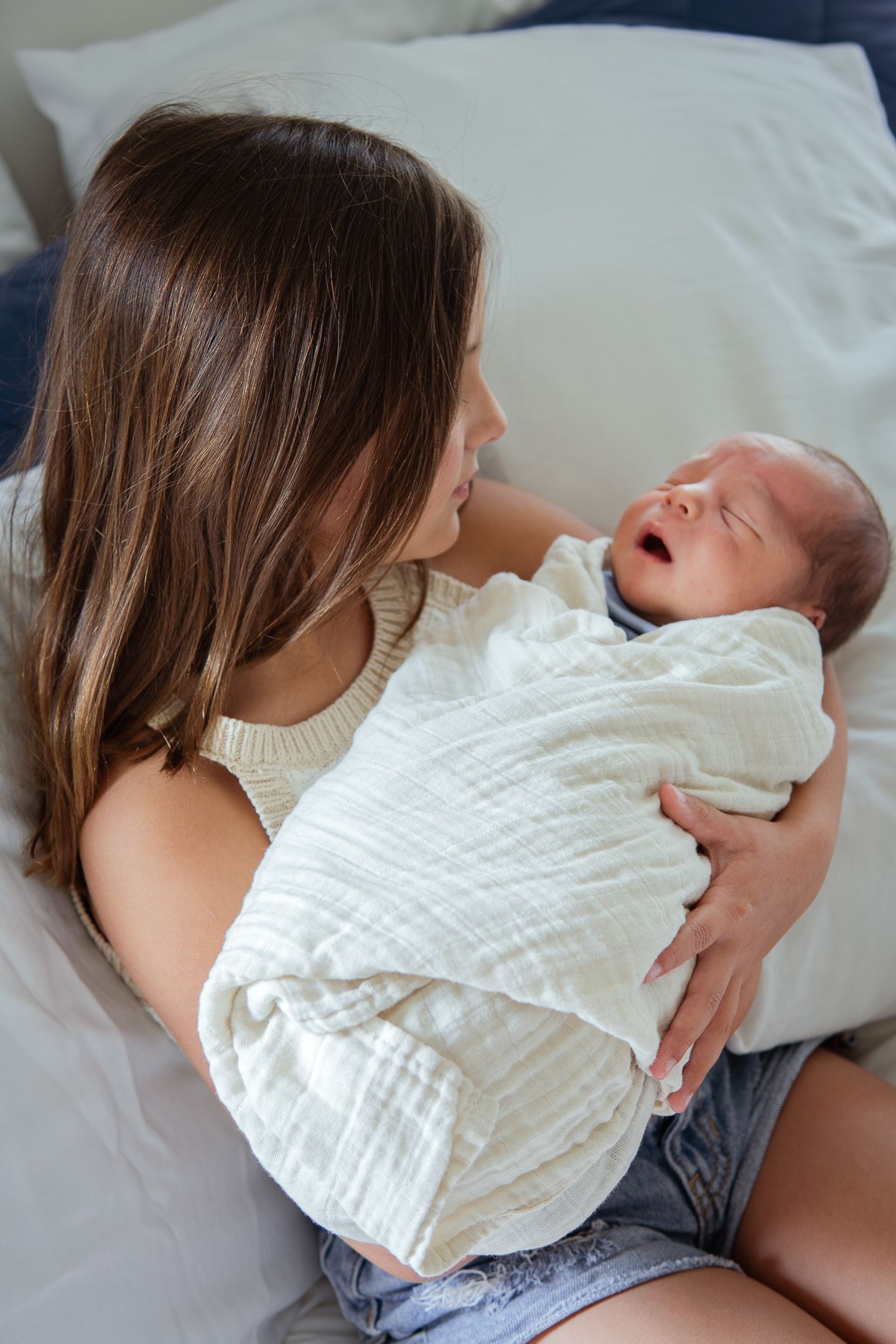 A newborn baby wrapped in white swaddle blanket crying while being held on a bed.