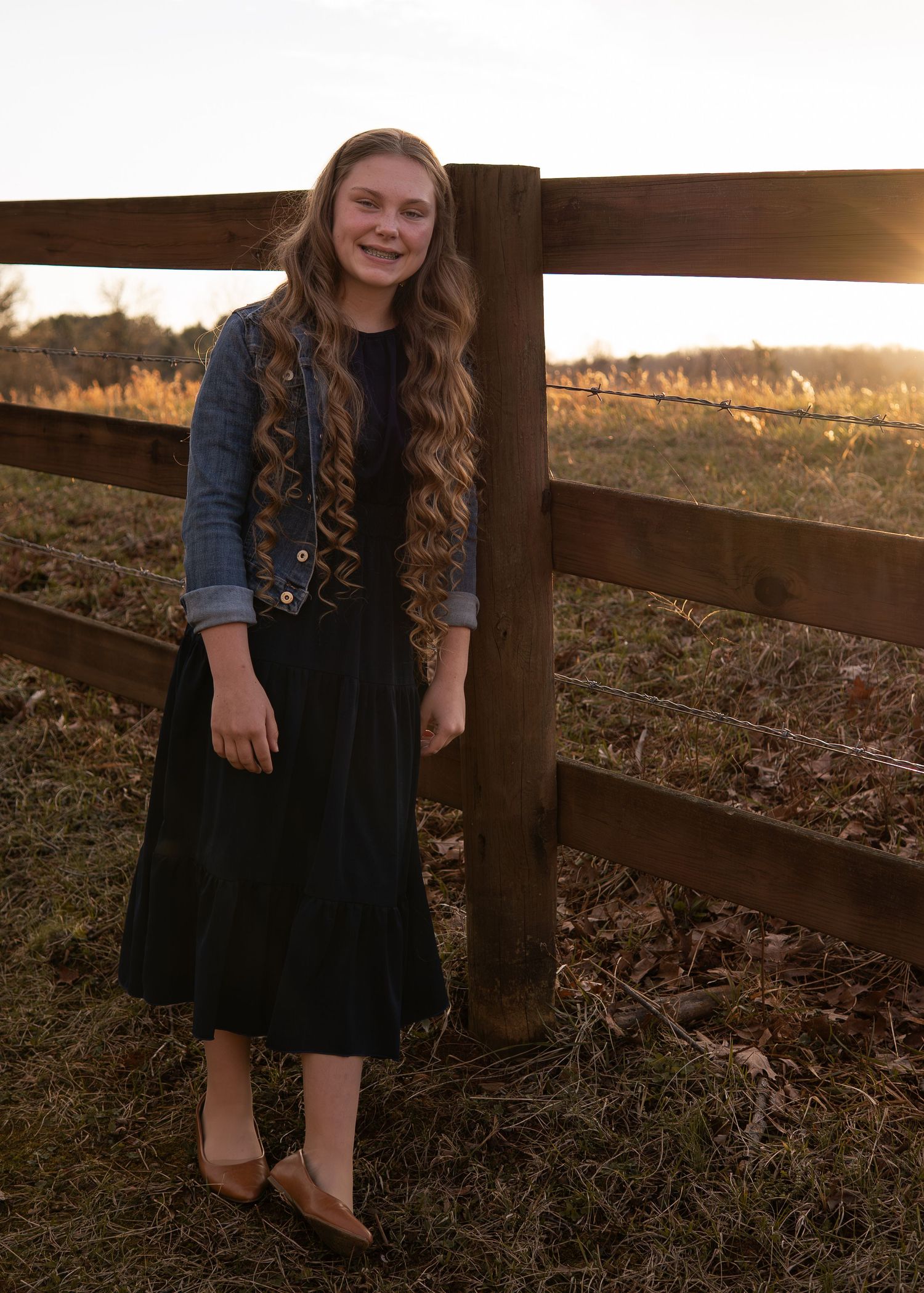 Teenage girl in black dress and denim vest posing by wooden fence at golden sunset.