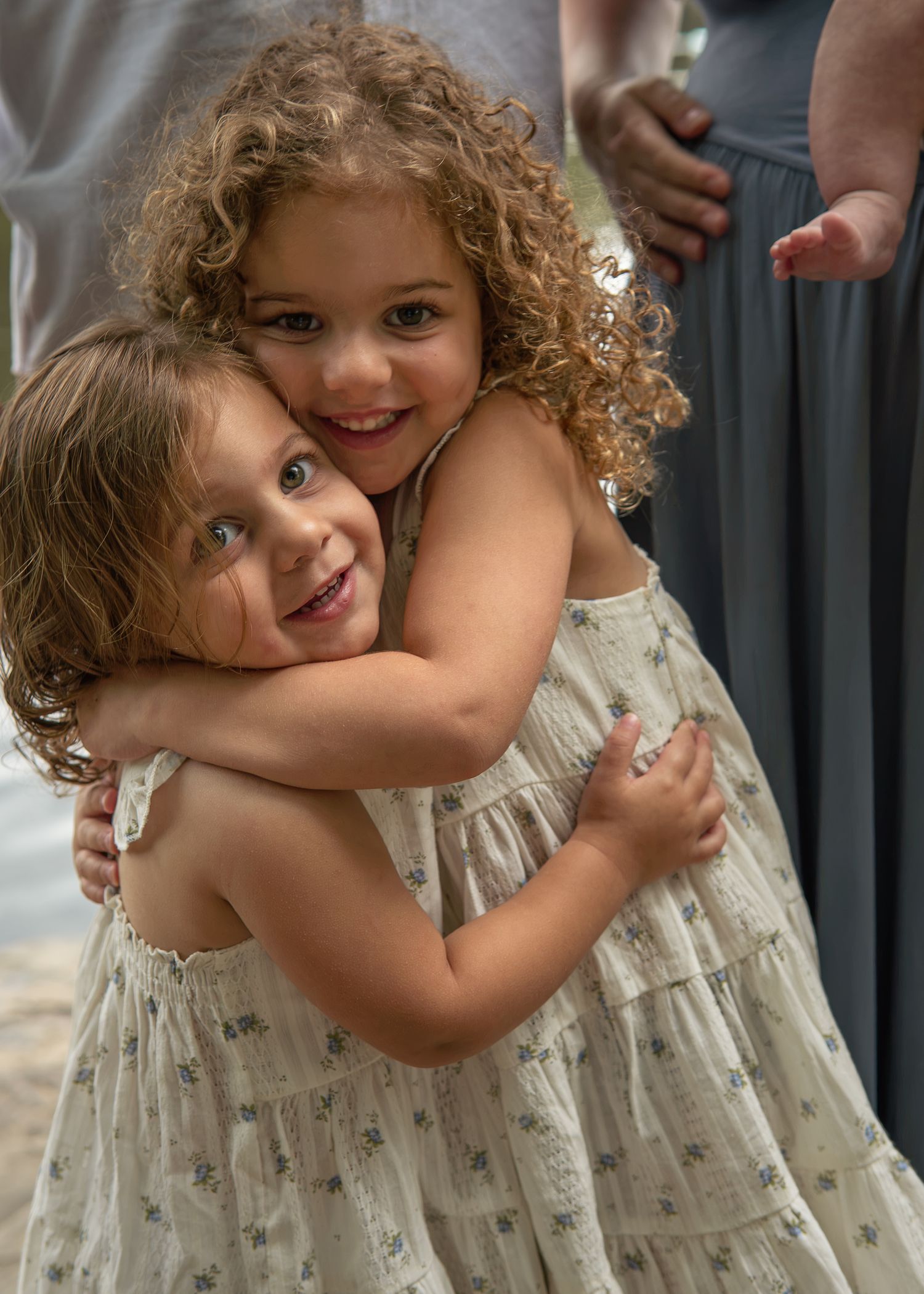 Two young girls with curly hair in floral dresses share a warm hug, smiling joyfully at an outdoor event.