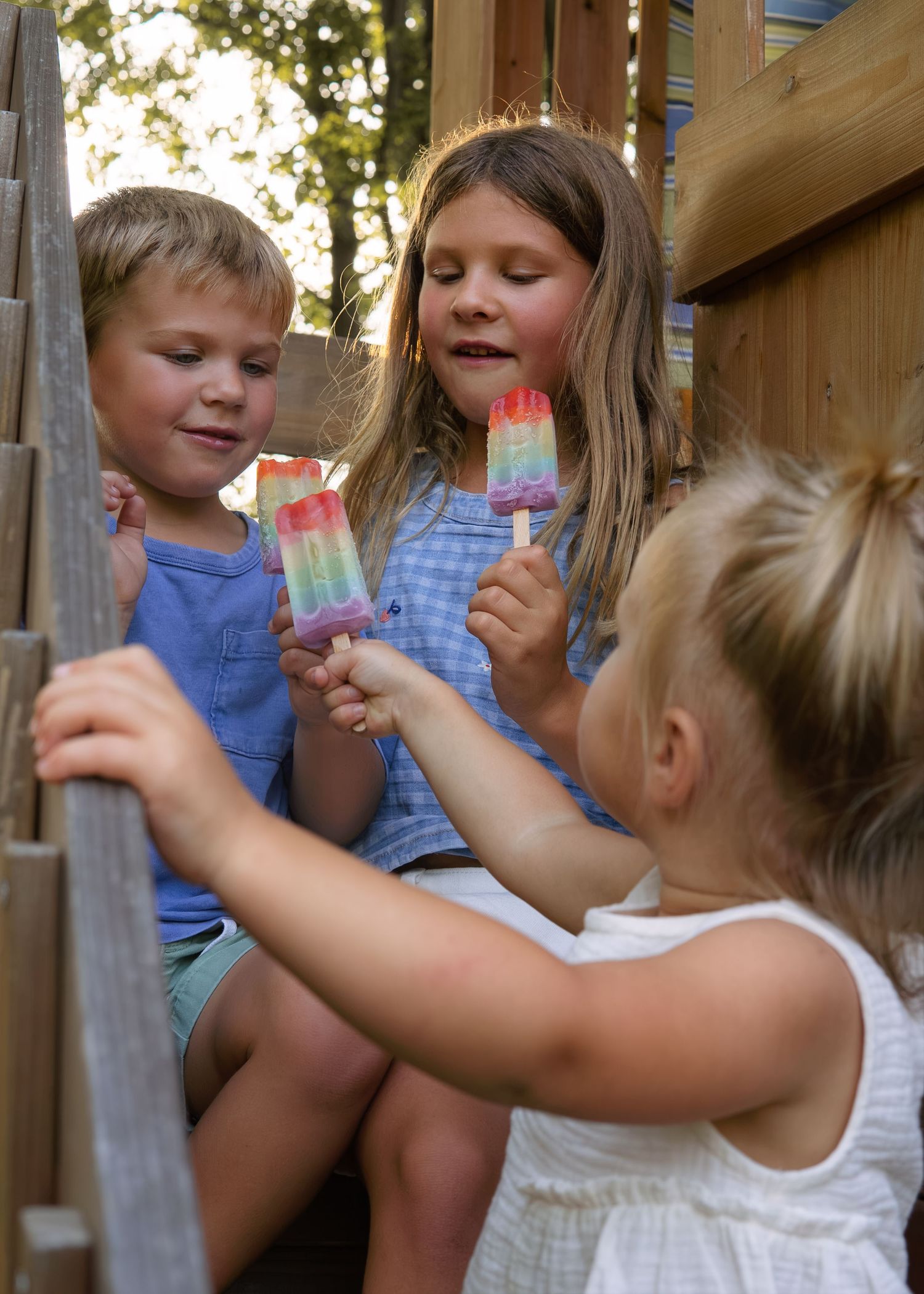Three children sitting on wooden play structure sharing colorful rainbow popsicles on a sunny summer day.