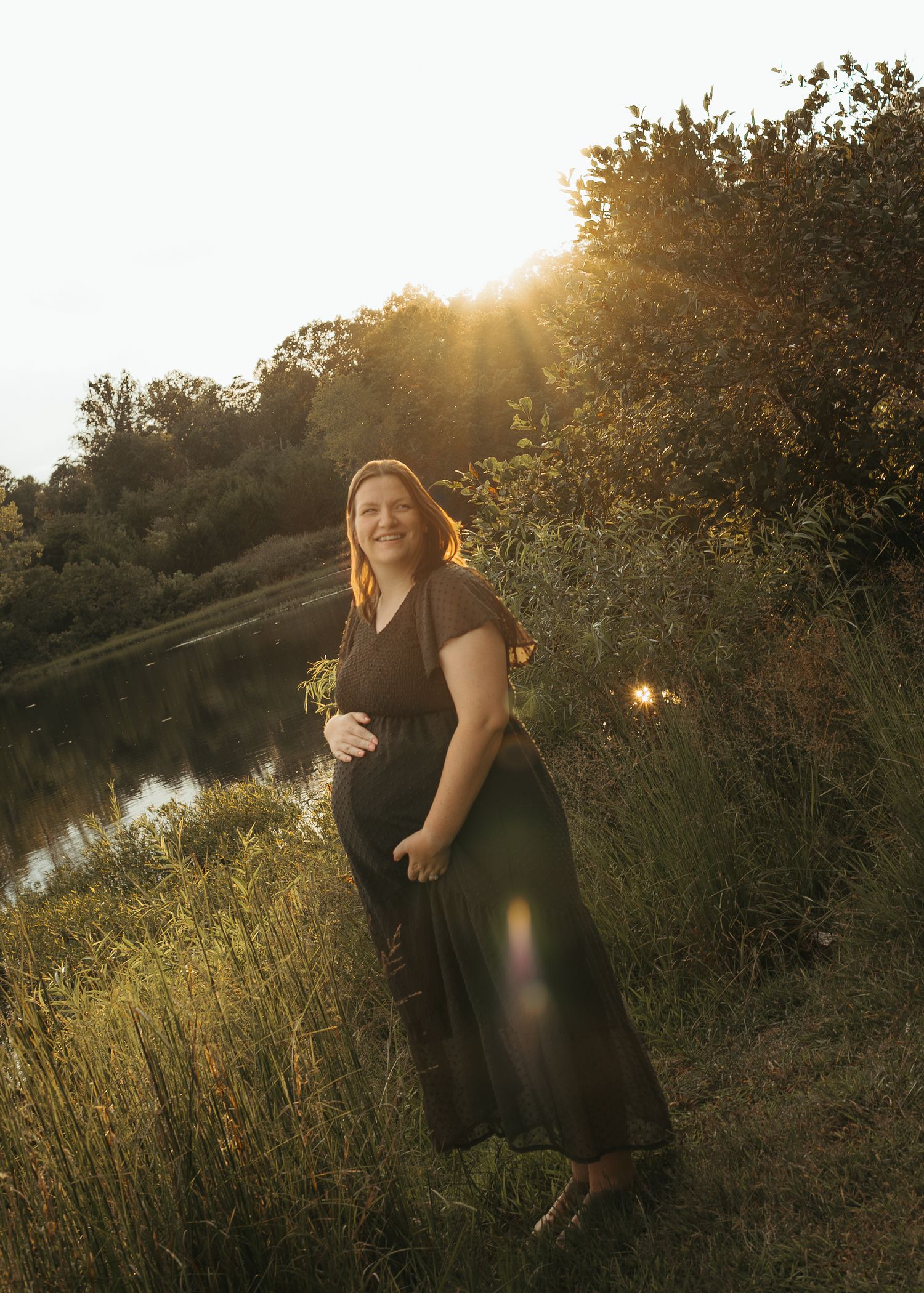Pregnant woman in dark olive maxi dress poses by a sunlit river at golden hour, smiling warmly.