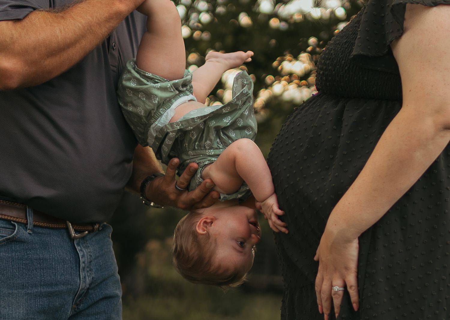 A father playfully holds a baby upside down outdoors while a pregnant mother stands nearby during a family photo session.
