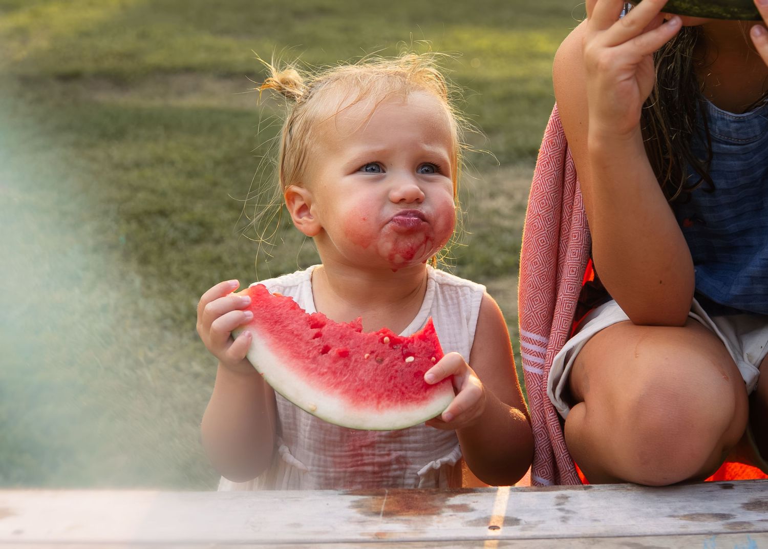 A cute toddler girl with pigtails enjoys a fresh slice of watermelon outdoors in summer.