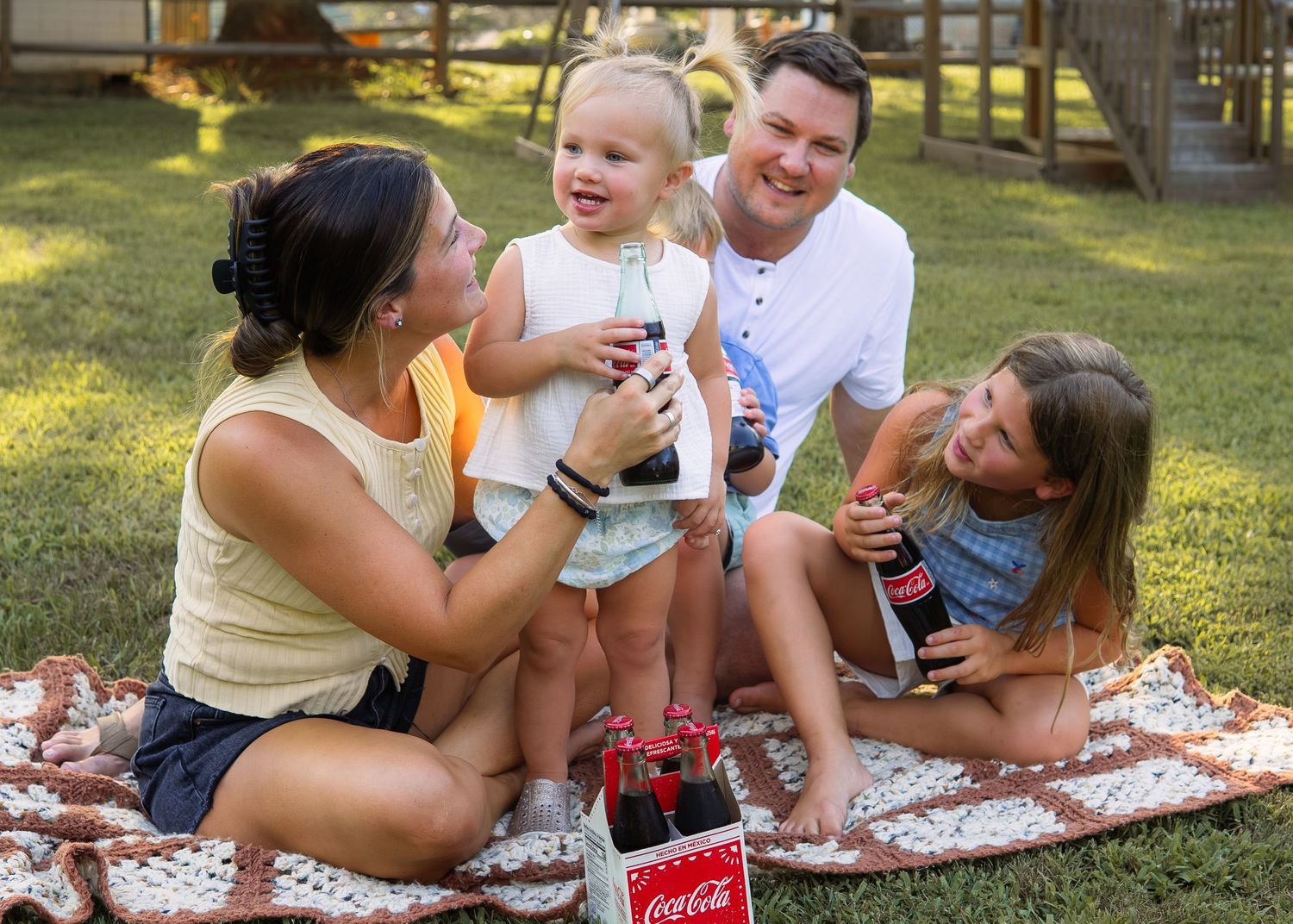 Family of four enjoying Coca-Cola bottles together on a picnic blanket outdoors on a sunny day.