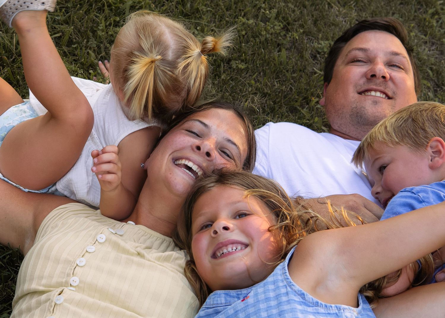 Happy family of five lying on green grass together, smiling and laughing outdoors on a sunny day.
